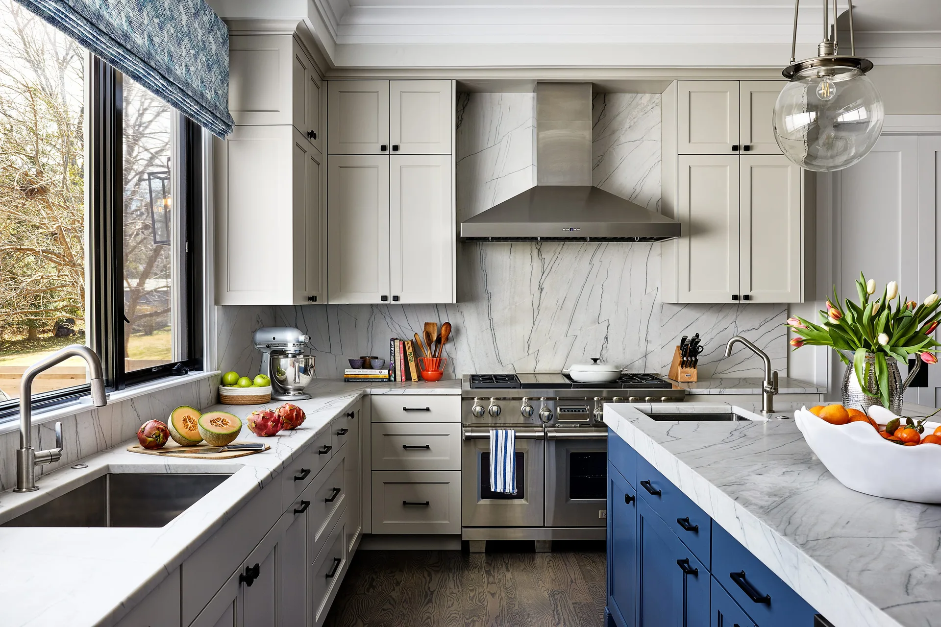 A transitional kitchen featuring light beige cabinets, a bold blue island with a marble countertop, and a matching full-height marble backsplash.