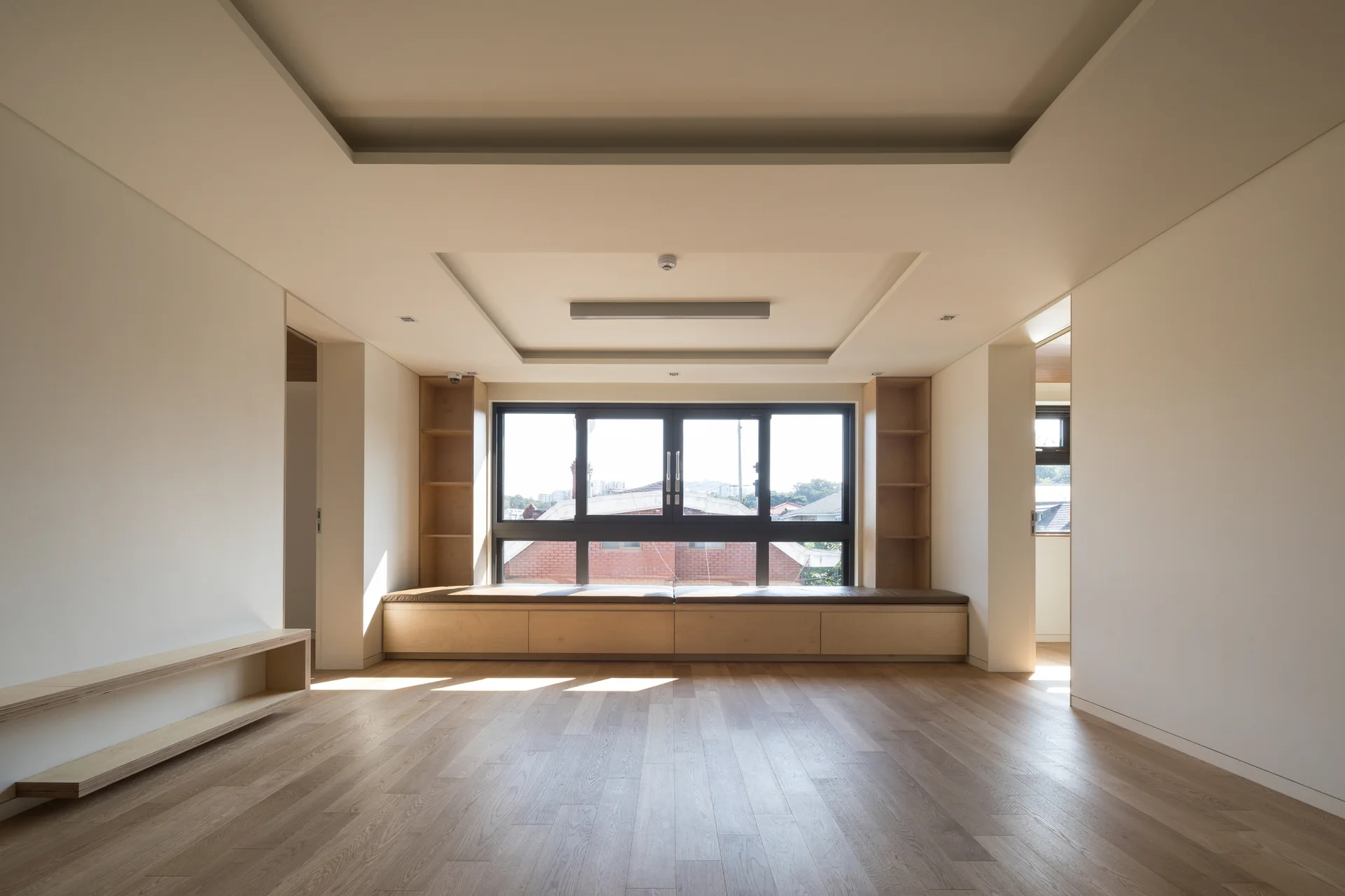 An empty minimalist room featuring light wood flooring, a built-in plywood window bench with shelving, and a tiered tray ceiling.