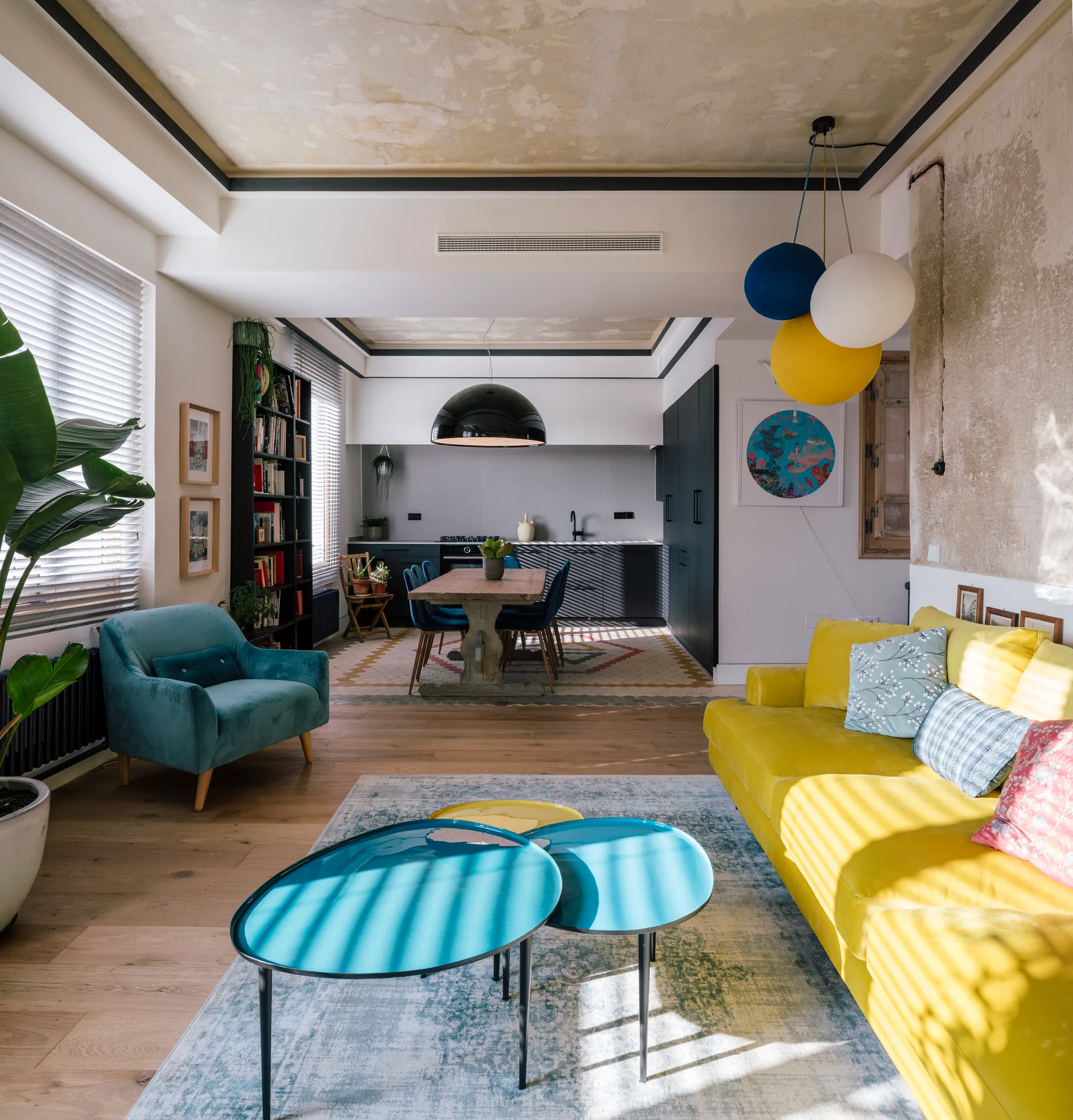 A bright open-concept living area with a yellow sofa, nested blue coffee tables, and a dining table set in the background.