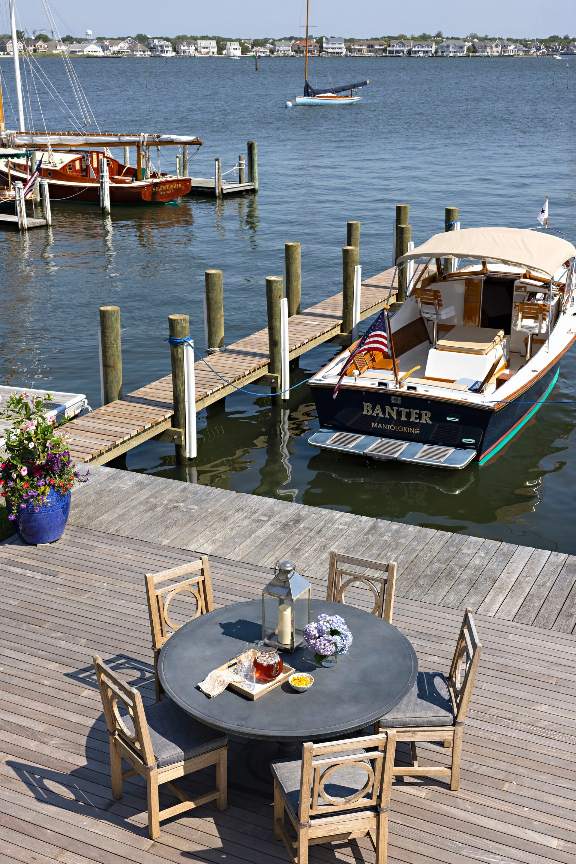 A round gray dining table with four wooden chairs sits on a weathered wooden dock overlooking a marina with moored boats.