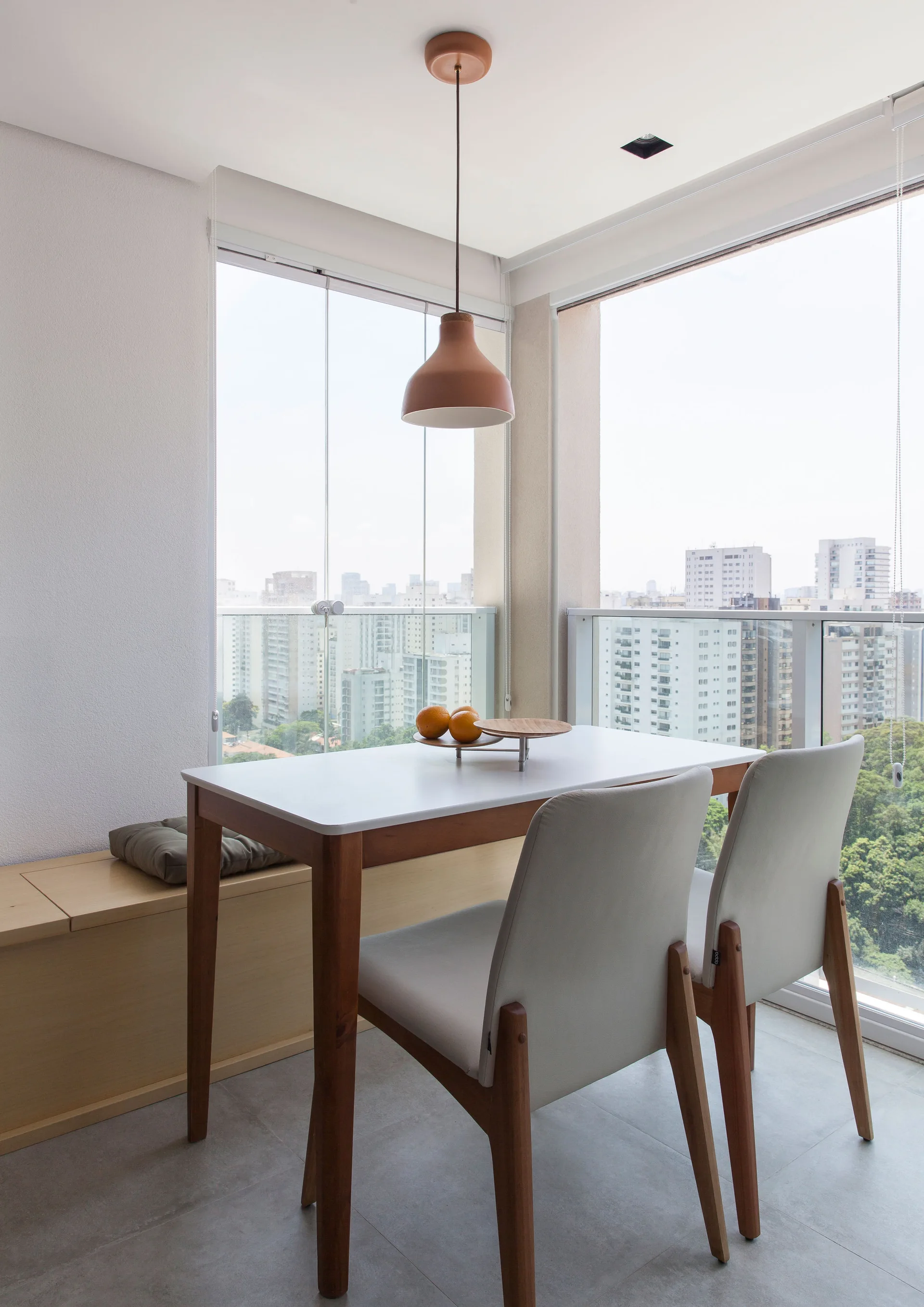 A small dining table and two grey chairs sit on an enclosed balcony next to a wooden bench and a large glass window overlooking a city.