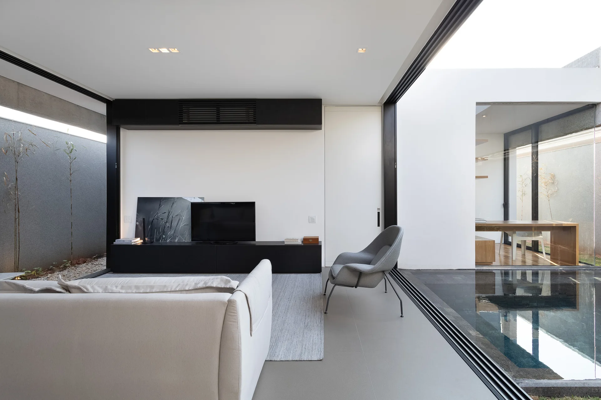 A minimalist living room features a white sofa, gray armchair, and low black console beside sliding glass doors opening to a courtyard pool.