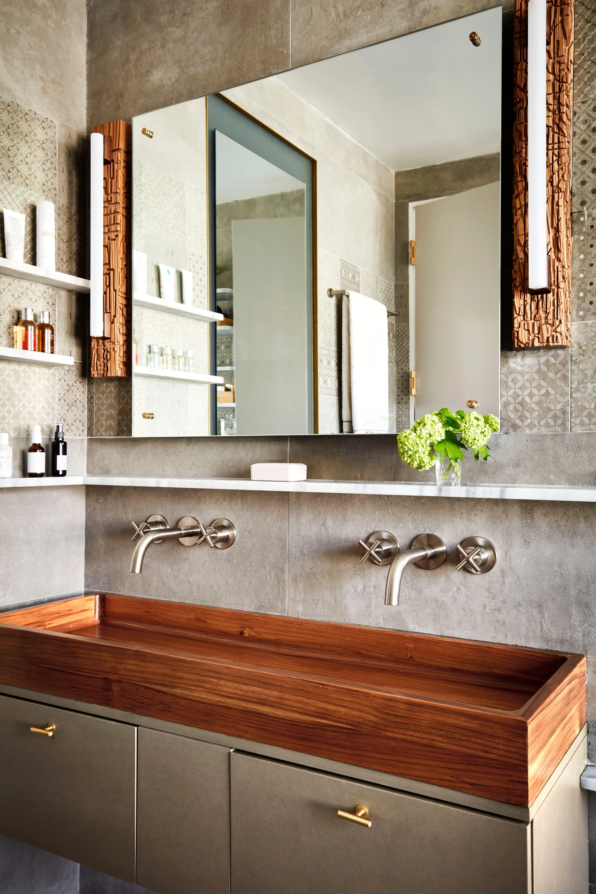 A modern bathroom vanity features a wood trough sink, wall-mounted faucets, a marble shelf, and a mirror with rustic wood sconces.