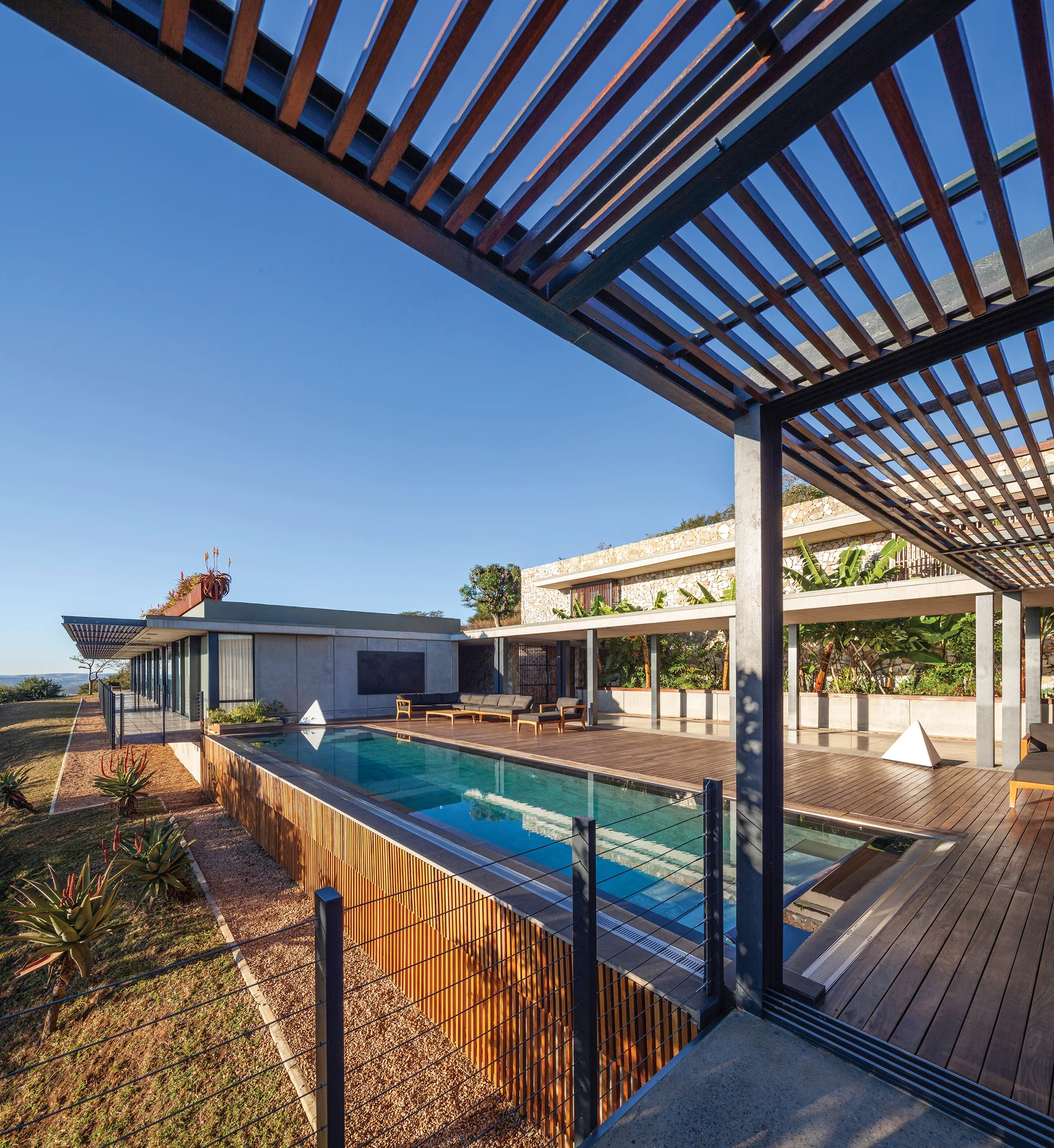 A modern rectangular swimming pool built into a wooden deck features a large overhead slatted pergola and outdoor lounge seating.
