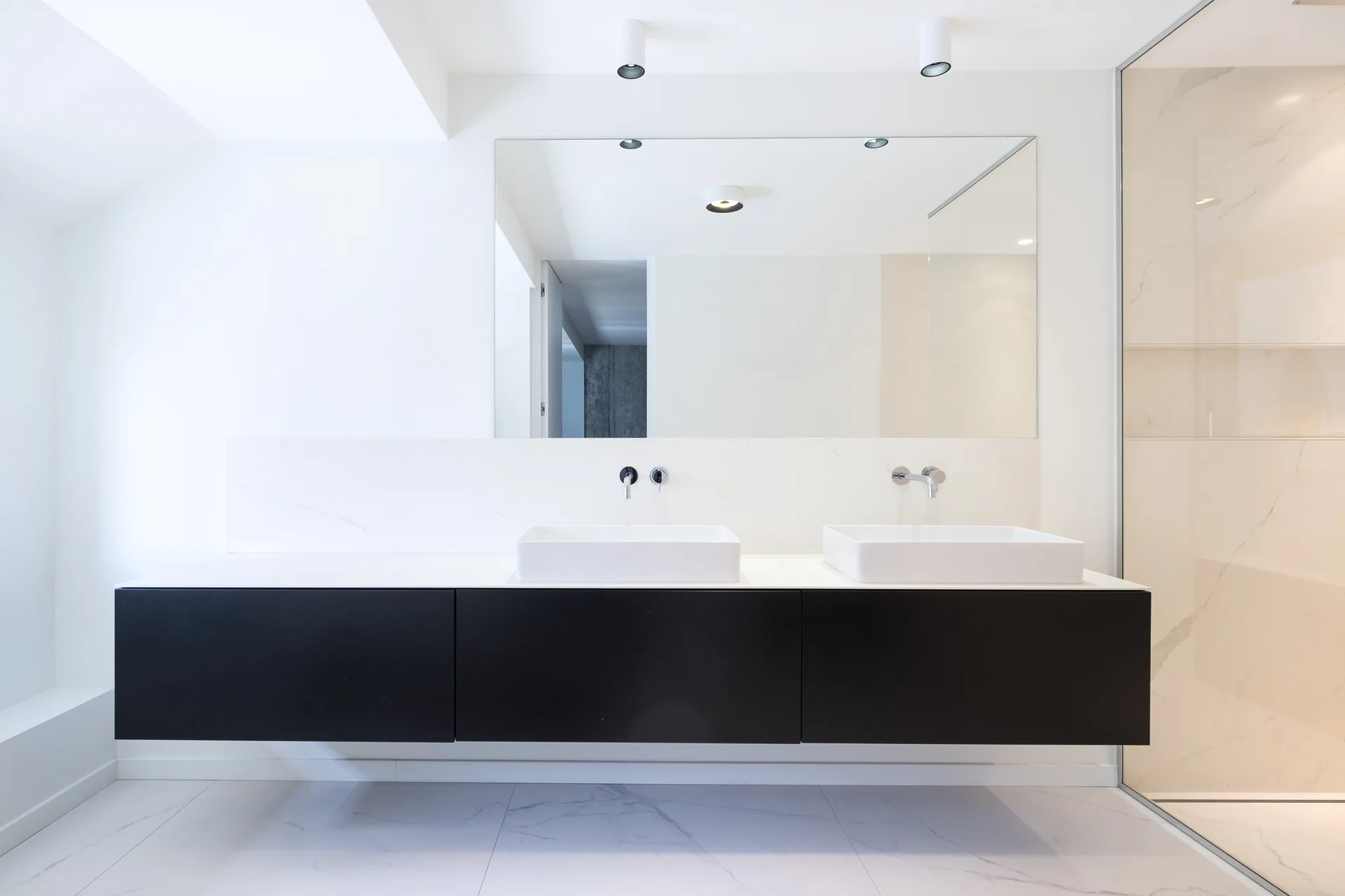 A minimalist bathroom featuring a black floating vanity, two white vessel sinks, and a large frameless mirror against white marble tiles.