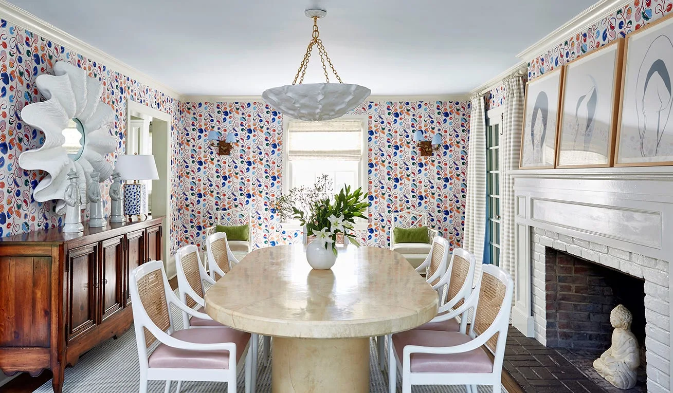 A dining room features an oval stone table surrounded by white cane-back chairs, set against colorful botanical wallpaper and a white brick fireplace.