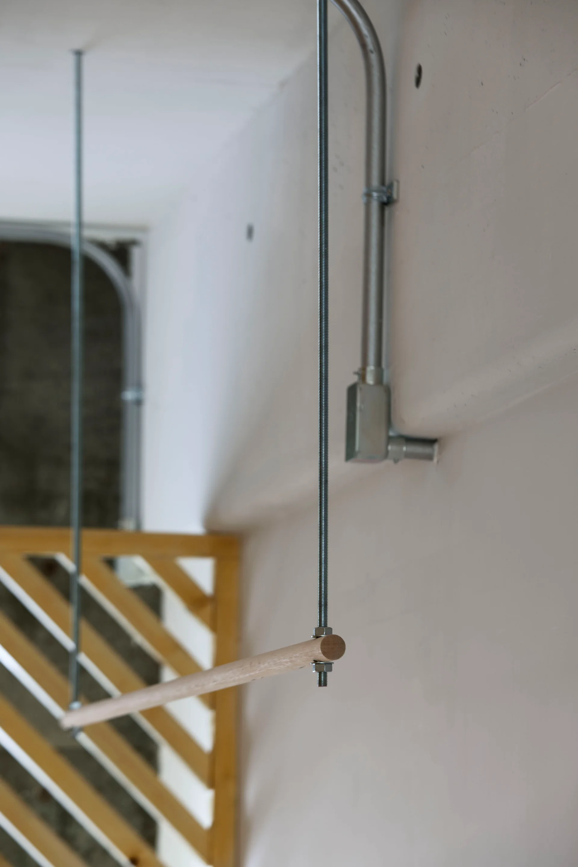 A wooden cylindrical handrail is suspended by thin threaded metal rods against an off-white wall with exposed metal conduit.
