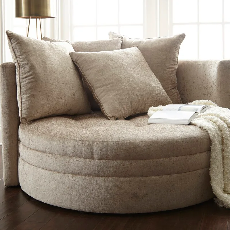 A large round beige upholstered armchair is piled with matching plush pillows, a white textured throw blanket, and an open book.