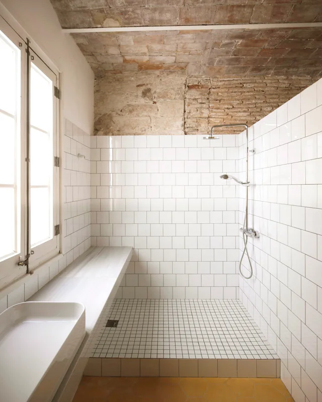 A minimalist bathroom with white tiled walls, an exposed brick ceiling, a yellow hexagonal tiled floor, and a long stone ledge.