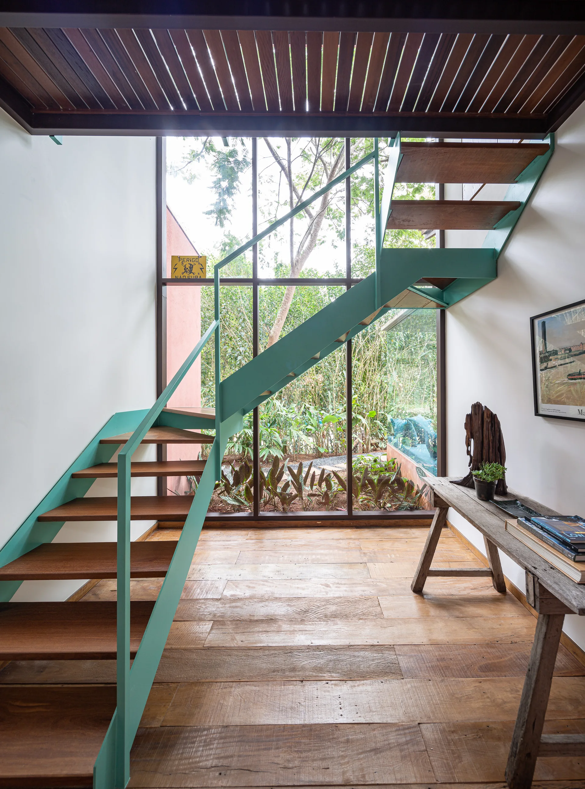 A modern teal metal staircase with wooden steps stands before a large floor-to-ceiling glass window overlooking a lush garden.