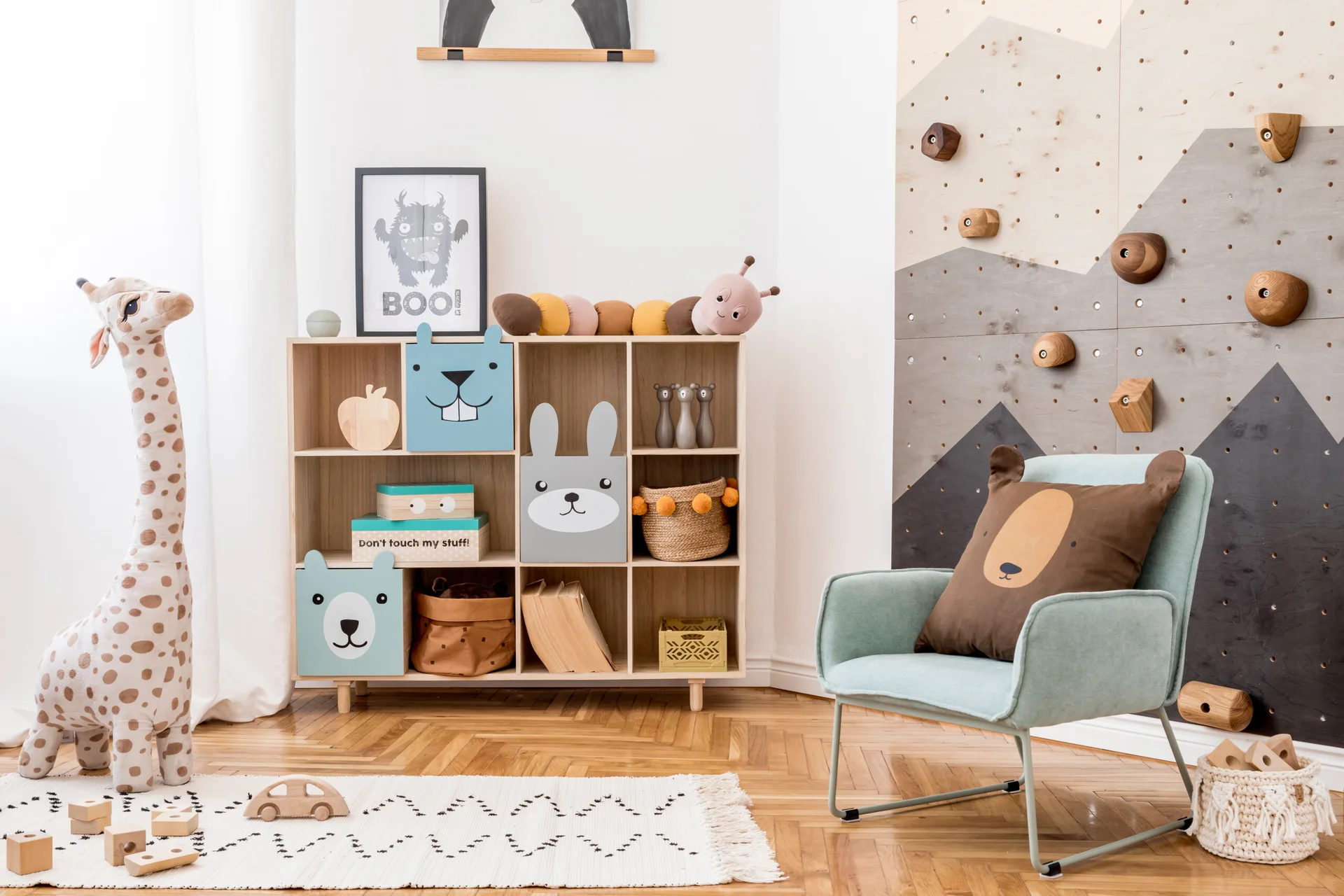 A children's playroom features a wooden cubby storage unit with animal faces, a mint green armchair, and a climbing wall with wooden holds.
