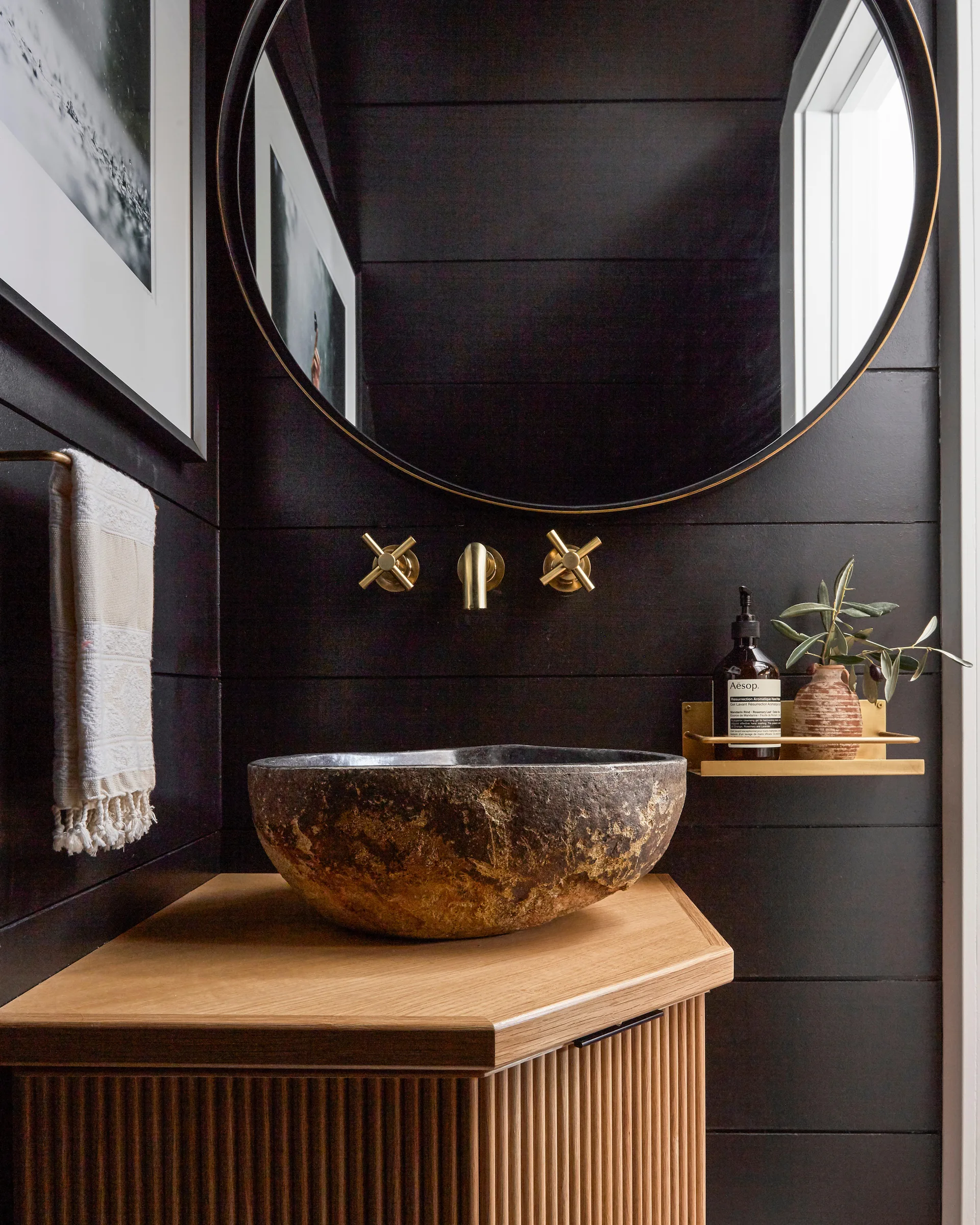 A black shiplap powder room features a rough stone vessel sink on a ribbed wood vanity with a round mirror and gold wall-mounted faucet.