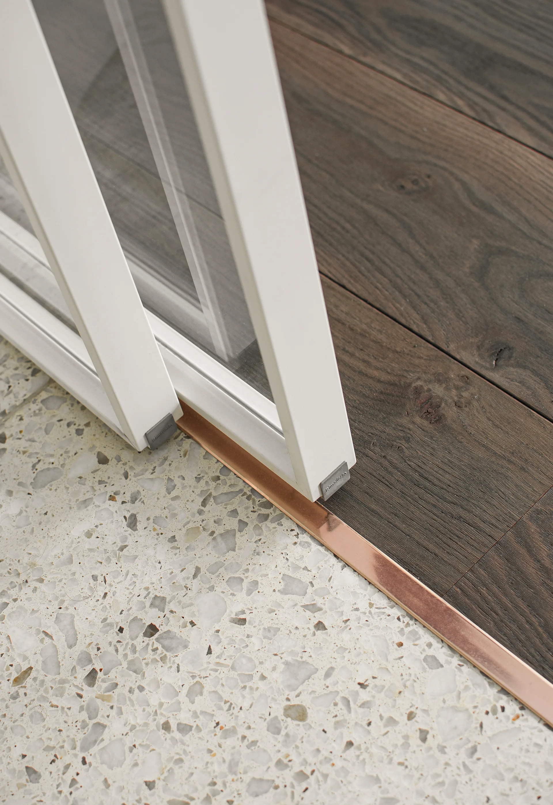 A close-up of a white sliding door track resting on a copper transition strip between light terrazzo and dark wood plank flooring.