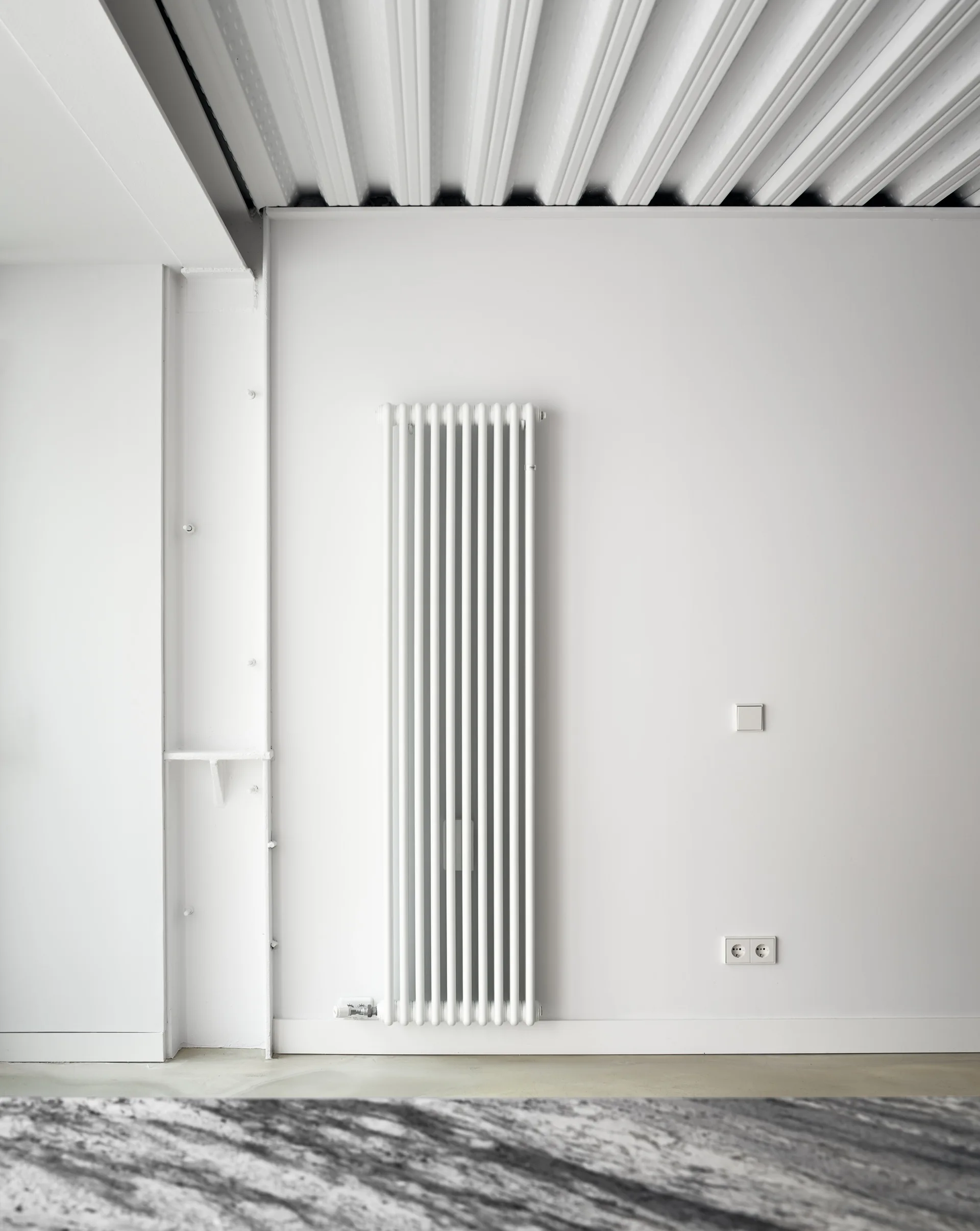 A tall white vertical radiator mounted on a flat white wall below a ribbed white industrial ceiling next to a structural column.