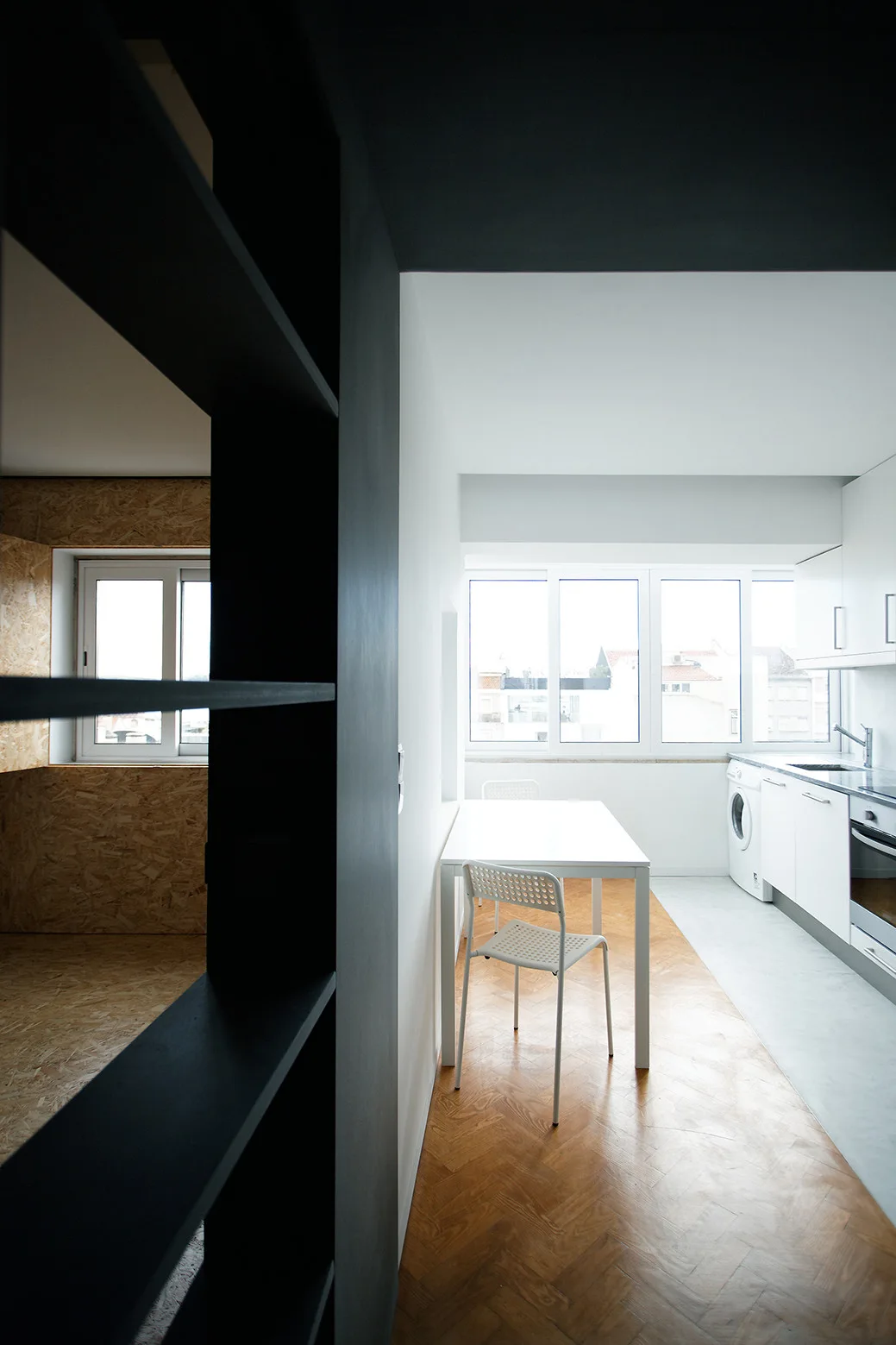 A minimalist white galley kitchen featuring a dining table with chairs, wood herringbone flooring, a black room divider, and an OSB paneled room.