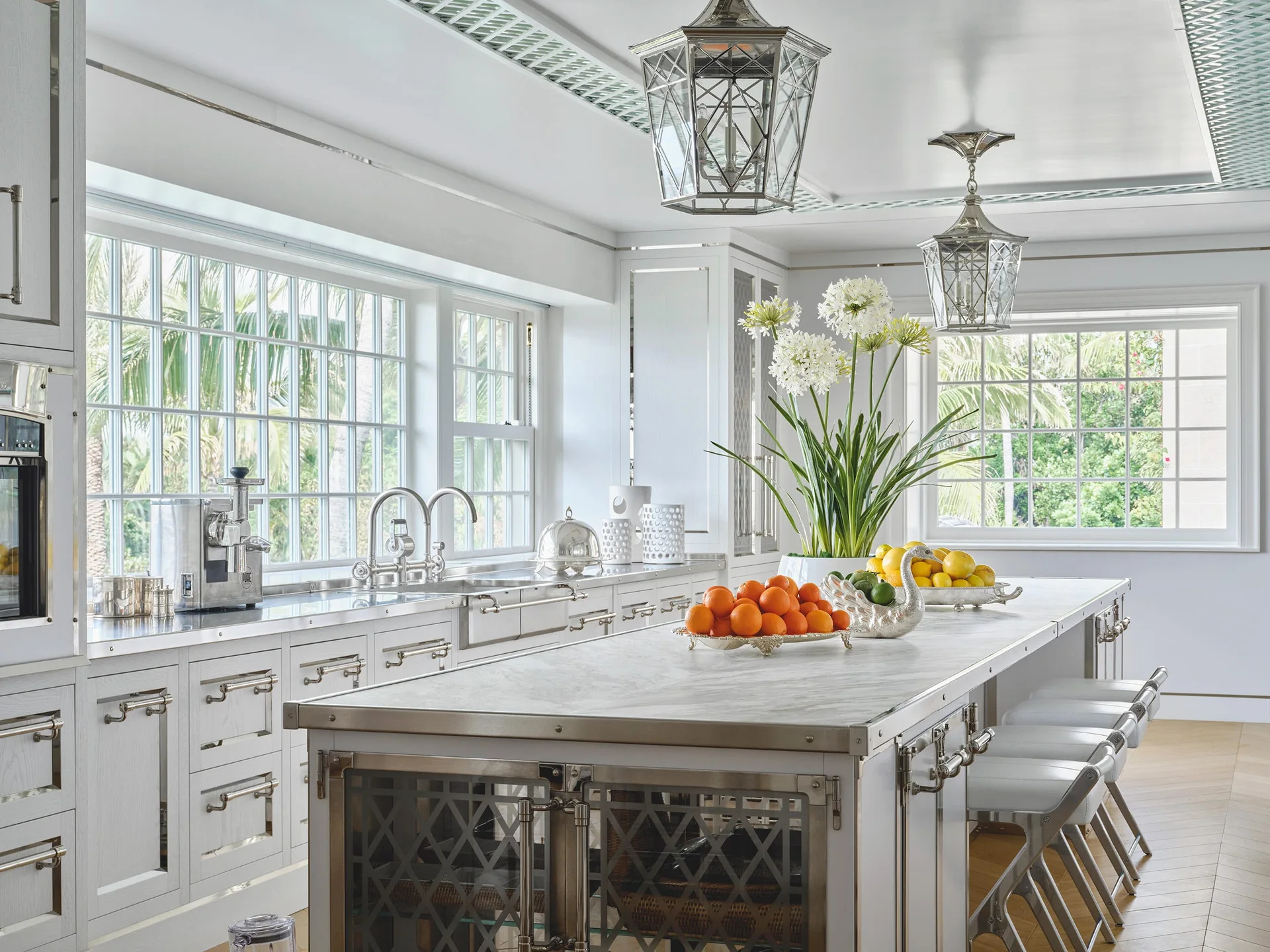 A bright white kitchen features a marble-topped central island with silver lattice detailing, matching metal pendant lanterns, and large grid windows.
