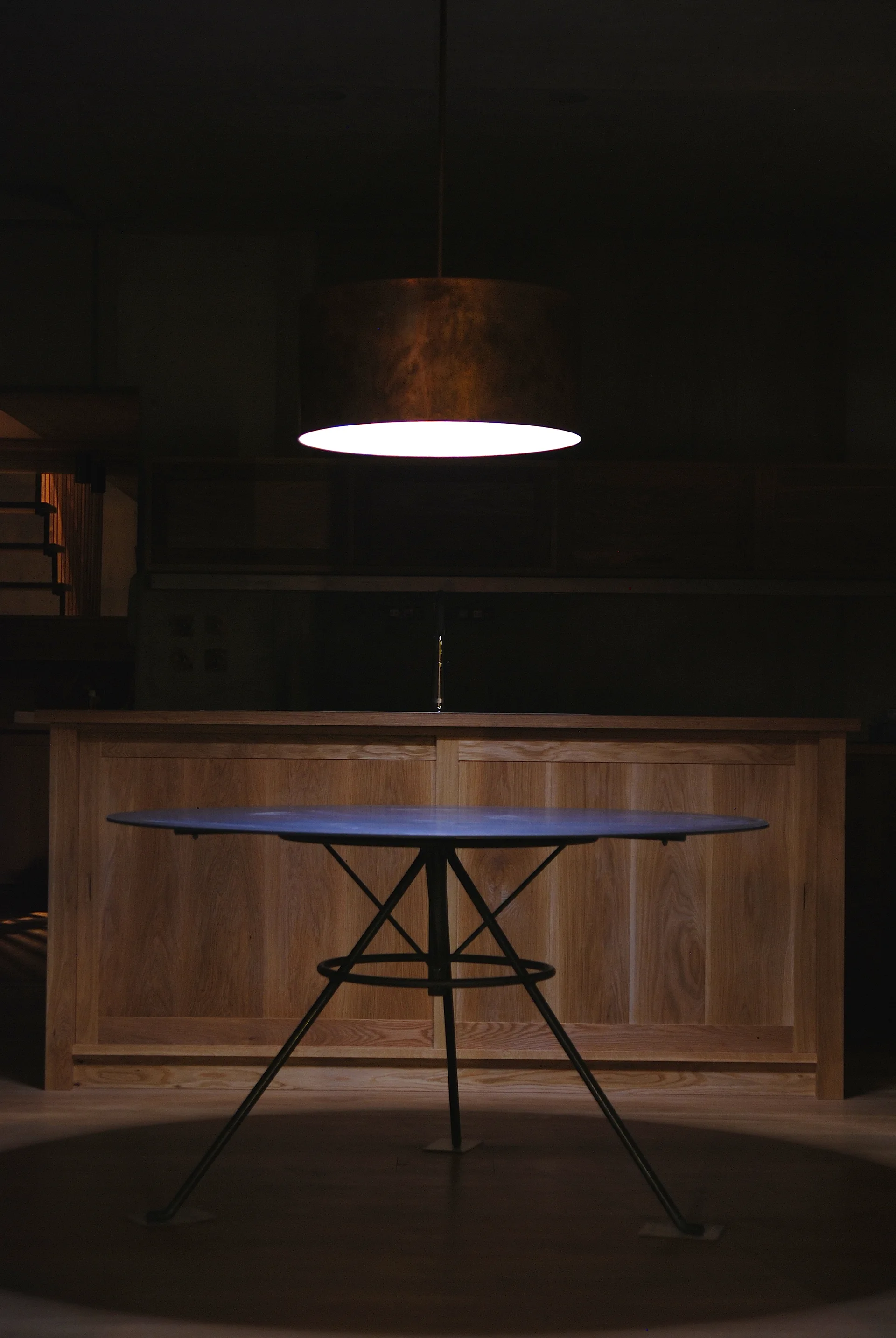 A circular dining table with metal legs sits under a large cylindrical brass pendant light in a dimly lit room with wood cabinetry.