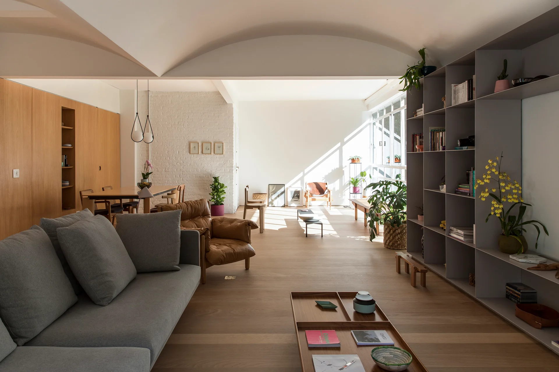 A minimalist open-plan living room features a grey sofa, light wood floors, a large gray shelving unit, and a unique white vaulted ceiling.
