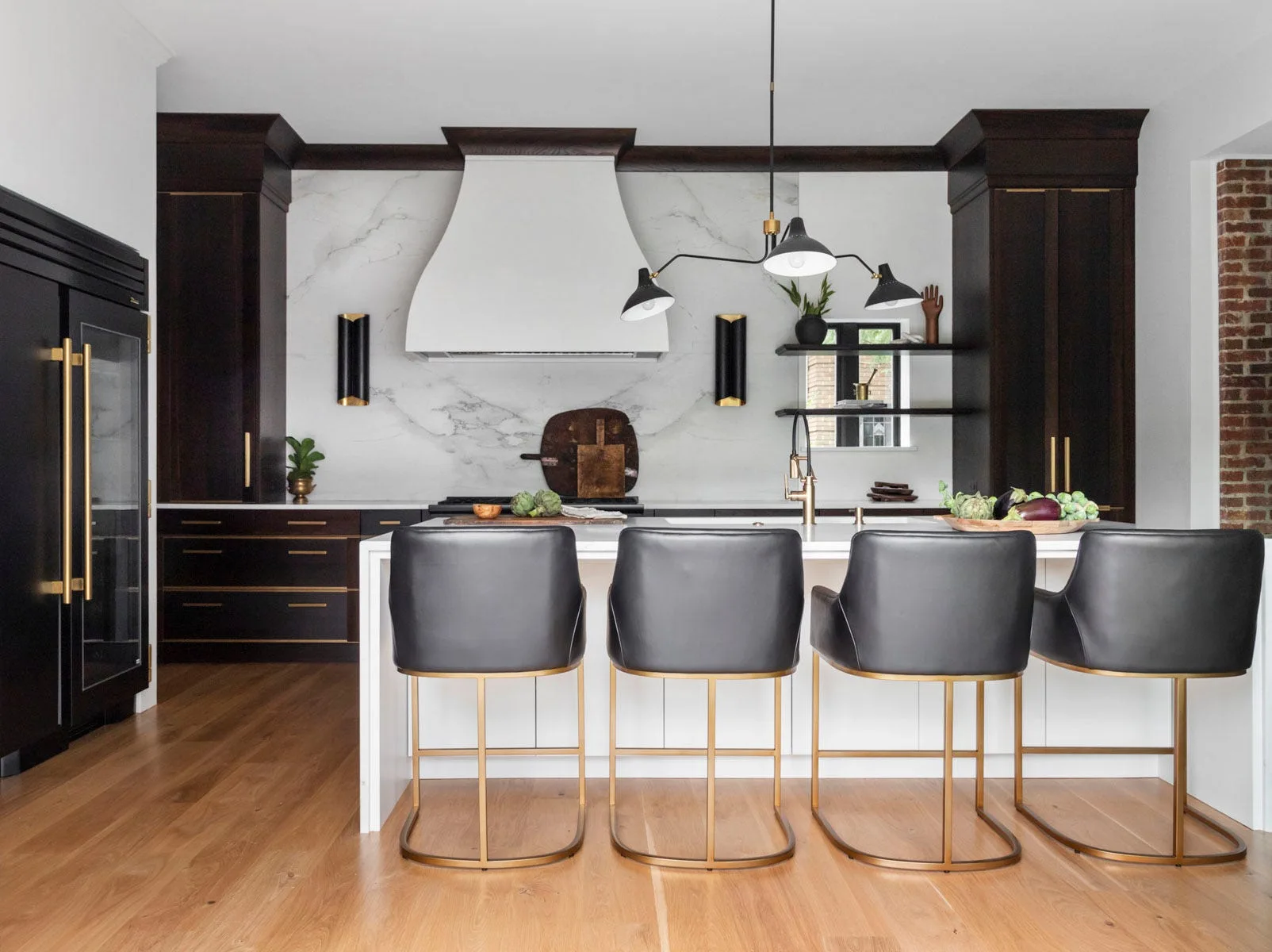 A modern kitchen features dark wood cabinetry, a white marble-look backsplash, a large white island, and four dark leather bar stools with gold frames.