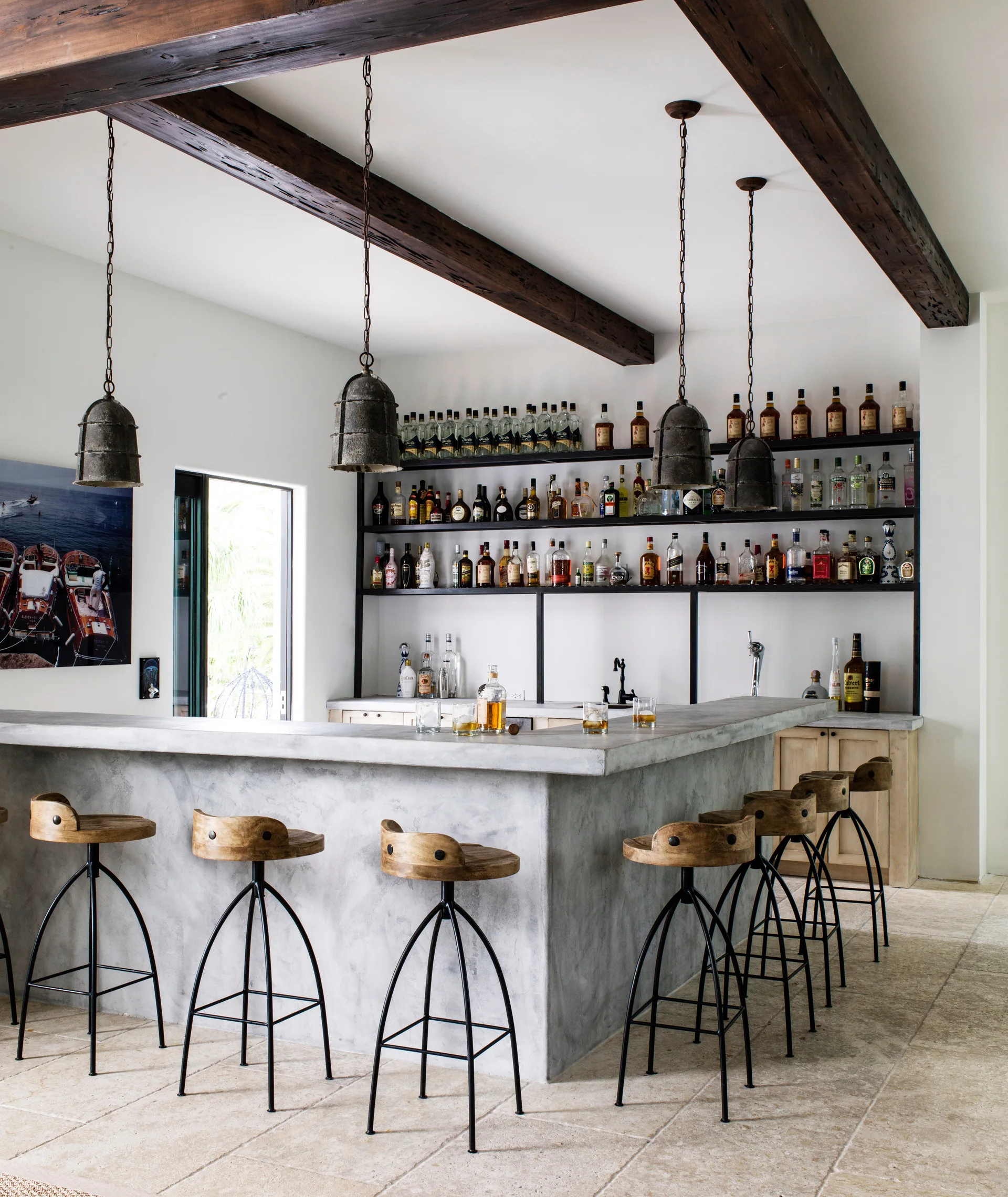 A home bar featuring a concrete counter, wooden swivel stools, open shelving for liquor bottles, and dark wooden ceiling beams.