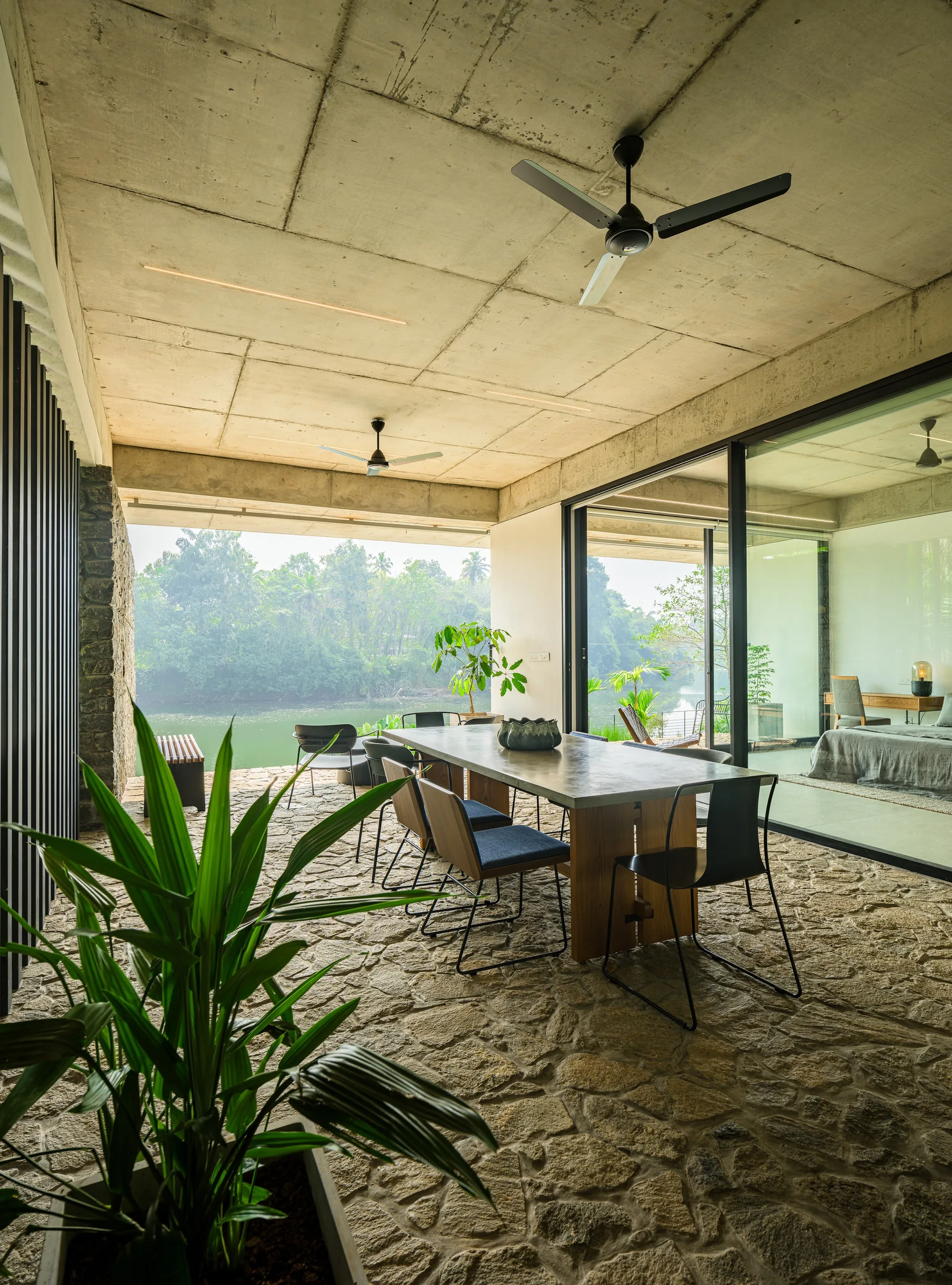 A covered outdoor dining area on a stone patio featuring a concrete table, modern chairs, and an exposed concrete ceiling overlooking a river.
