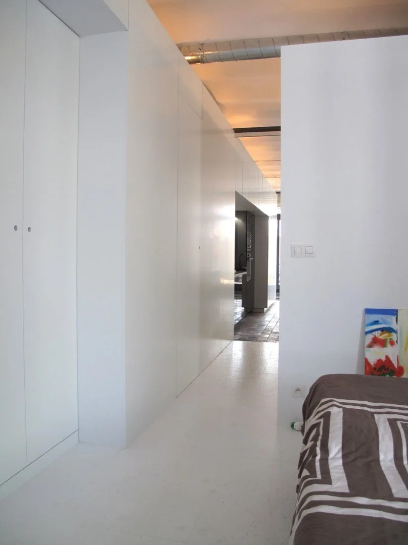 A minimalist white bedroom hallway featuring seamless floor-to-ceiling cabinets, a white floor, and a bed with brown patterned bedding.