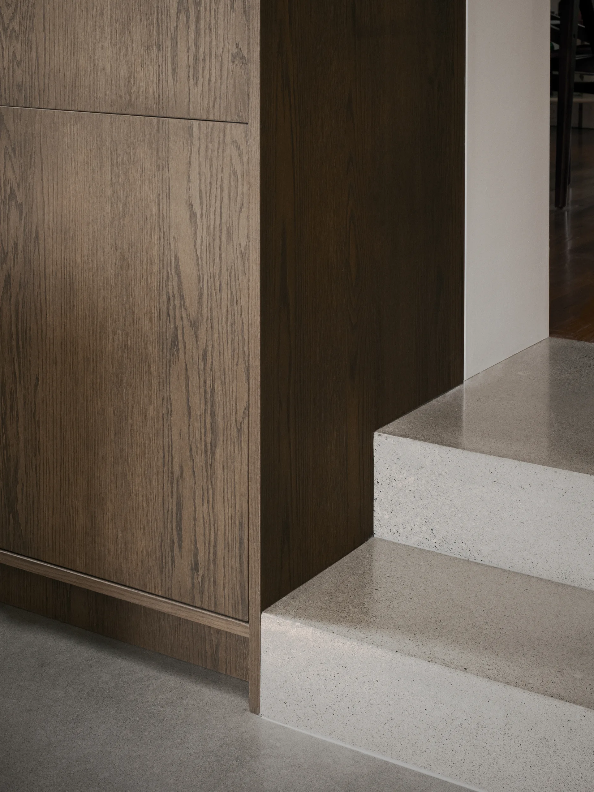 Polished concrete steps lead up beside a large dark wood cabinet with a visible grain pattern in a minimalist interior.