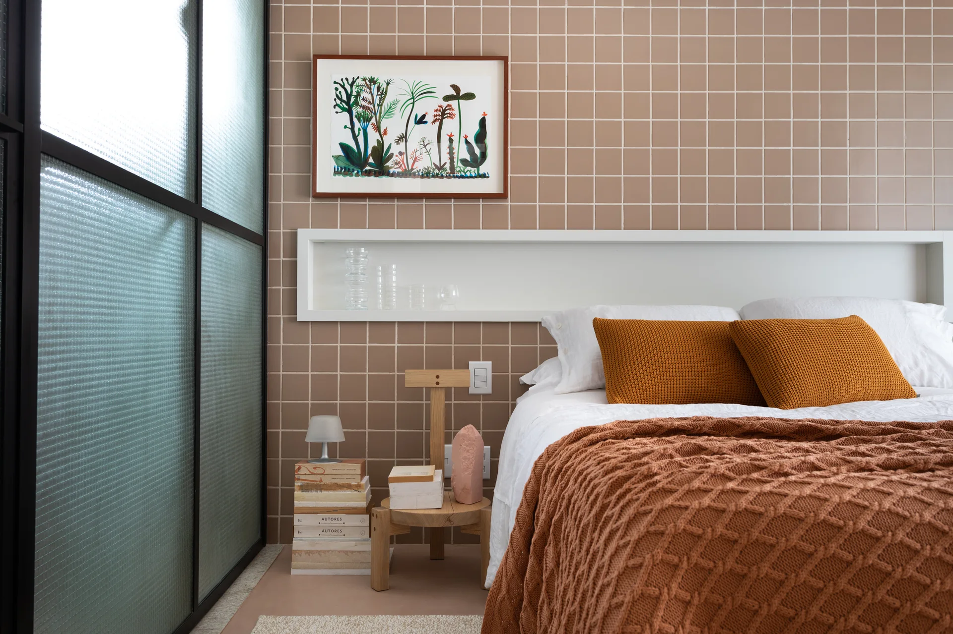 A bed with textured orange and brown blankets sits against a brown tiled wall with a built-in horizontal white shelf.