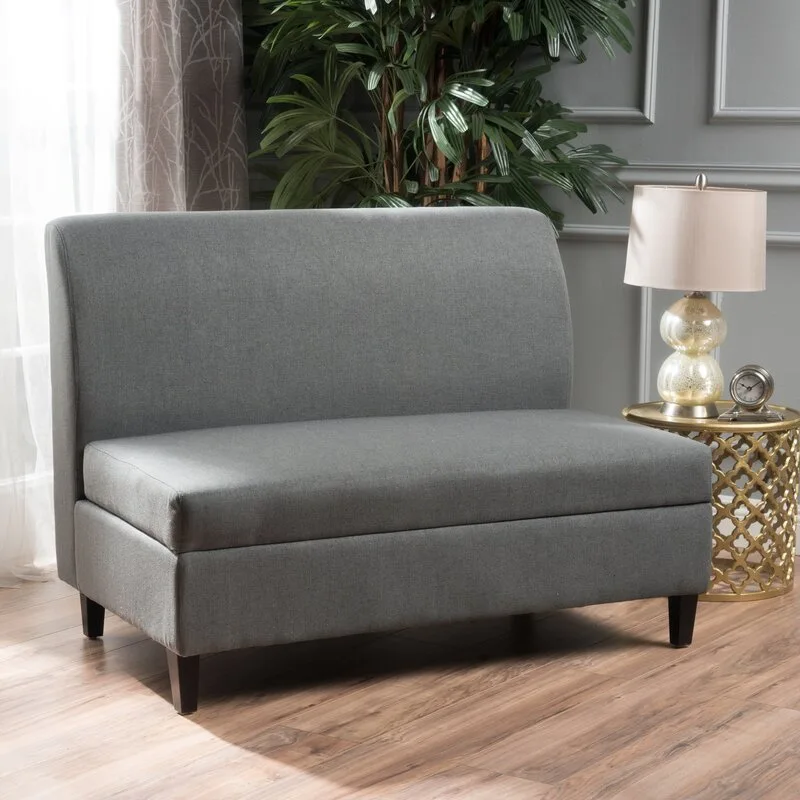 A gray upholstered armless loveseat sits next to a gold lattice side table with a table lamp in a room with gray paneled walls.
