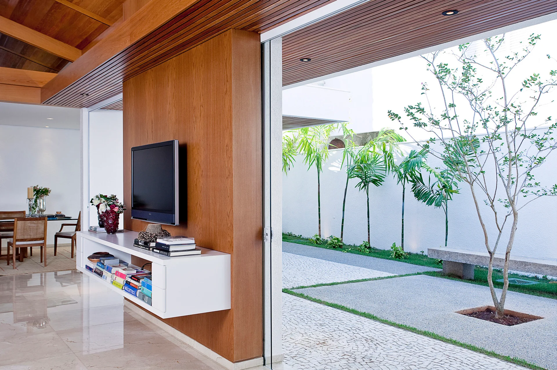 A contemporary living room features a wood accent wall with a mounted television and white floating console next to a sliding glass door opening to a courtyard.