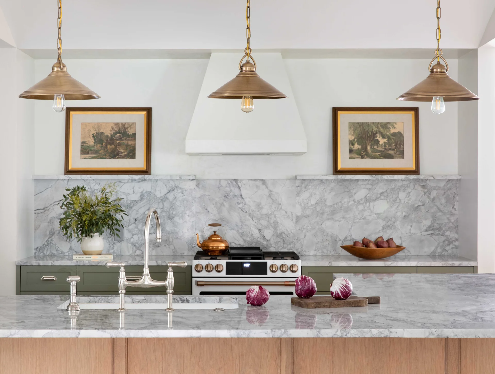 A kitchen features a marble-topped island with a sink, olive-green lower cabinets, a marble slab backsplash, and three brass pendant lights hanging above.