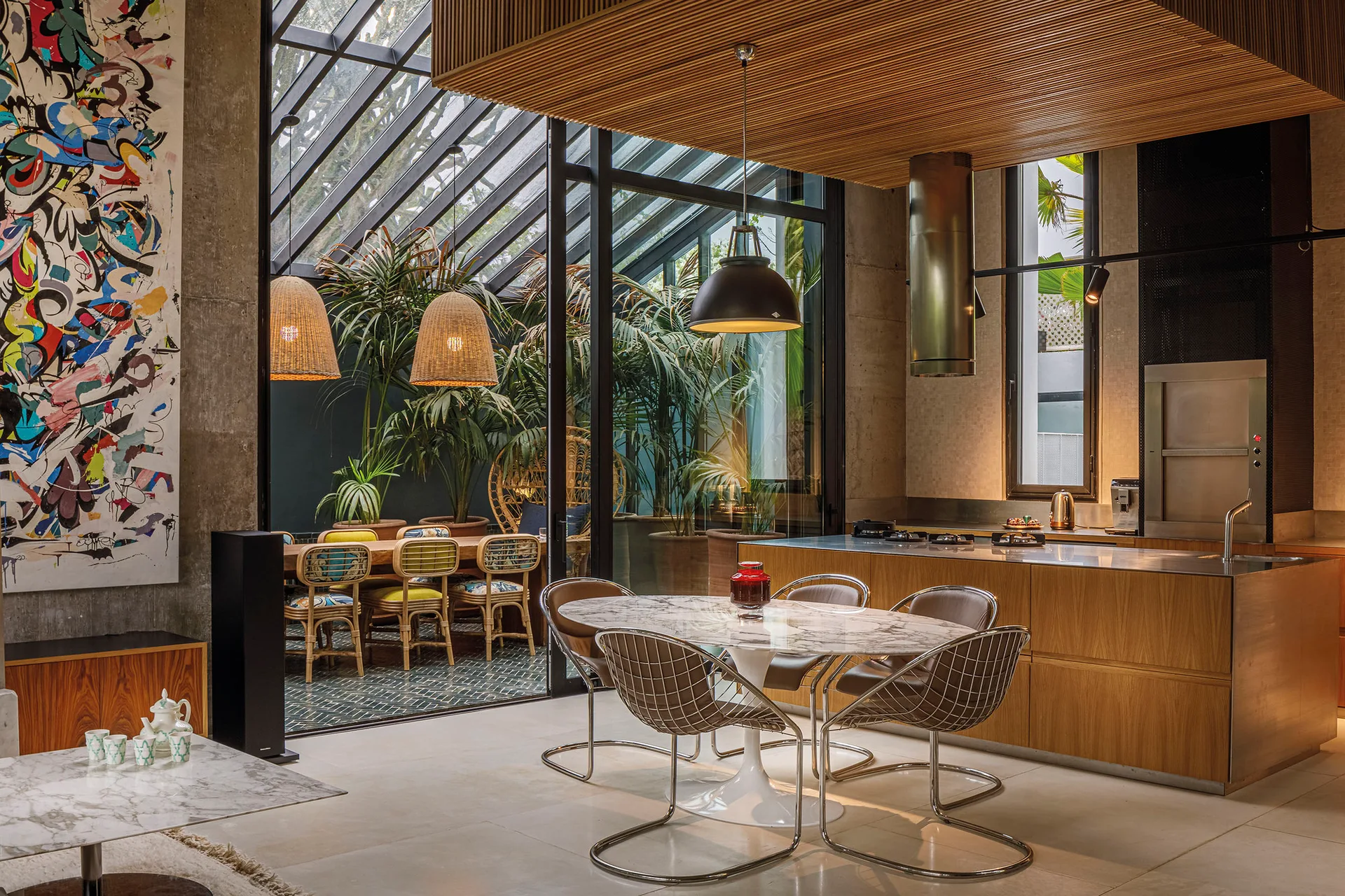 A modern dining room features a marble-topped pedestal table, wire mesh chairs, a slatted wood ceiling, and an indoor greenhouse visible through glass walls.