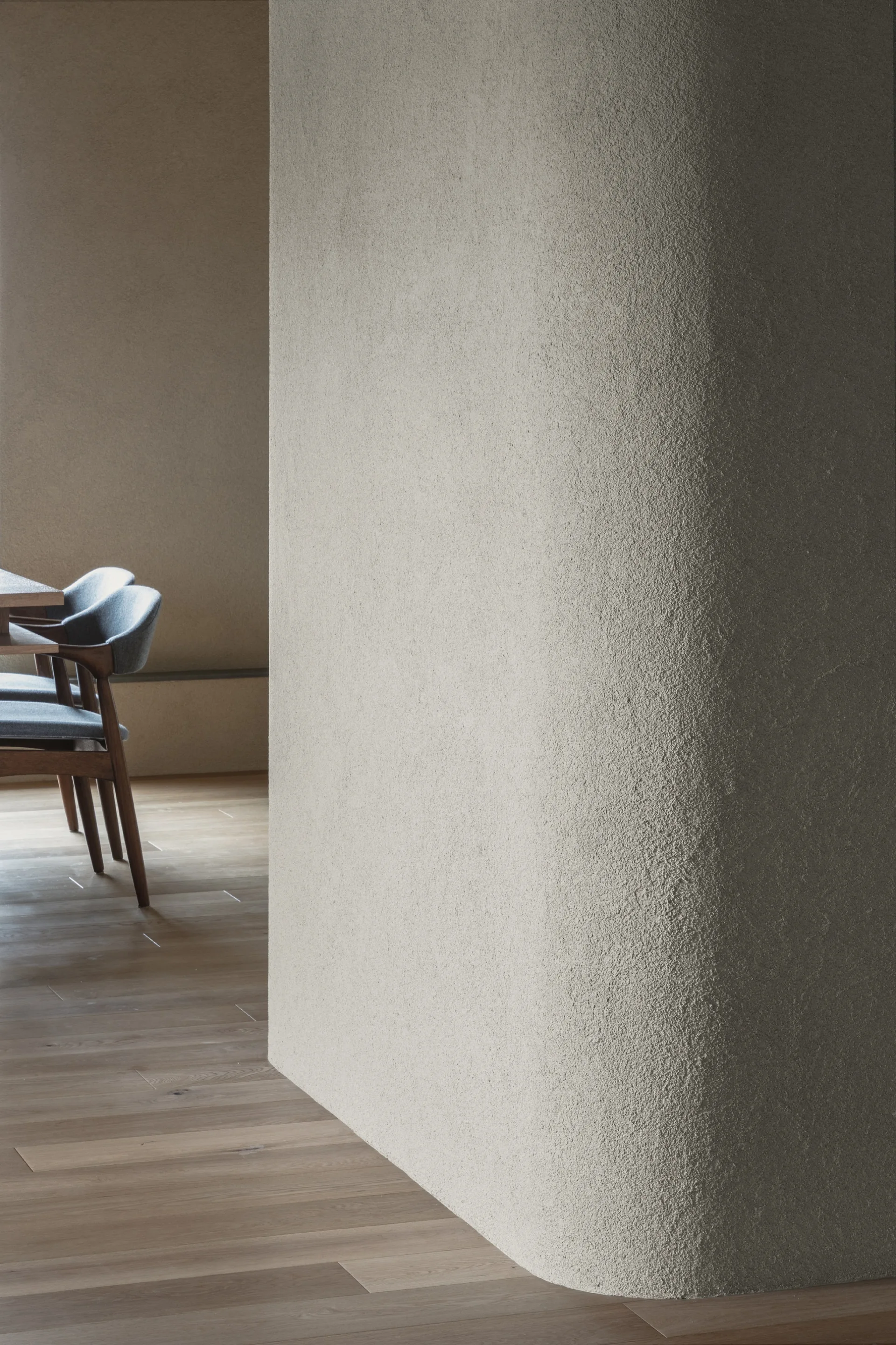 A curved textured plaster wall partition stands next to a wooden dining set with upholstered chairs on light hardwood flooring.