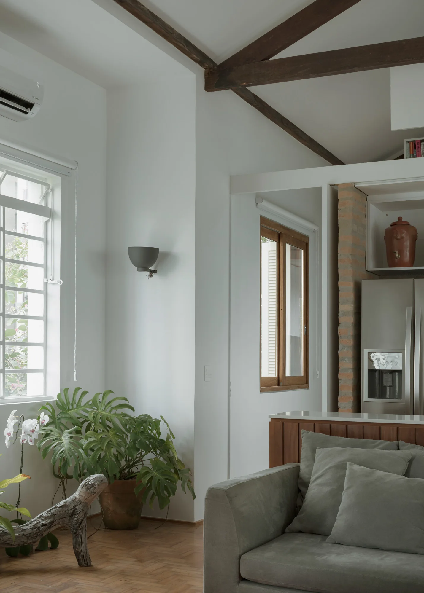 A living room featuring a grey upholstered sofa, exposed wooden ceiling beams, a herringbone wood floor, and a large monstera plant by the window.
