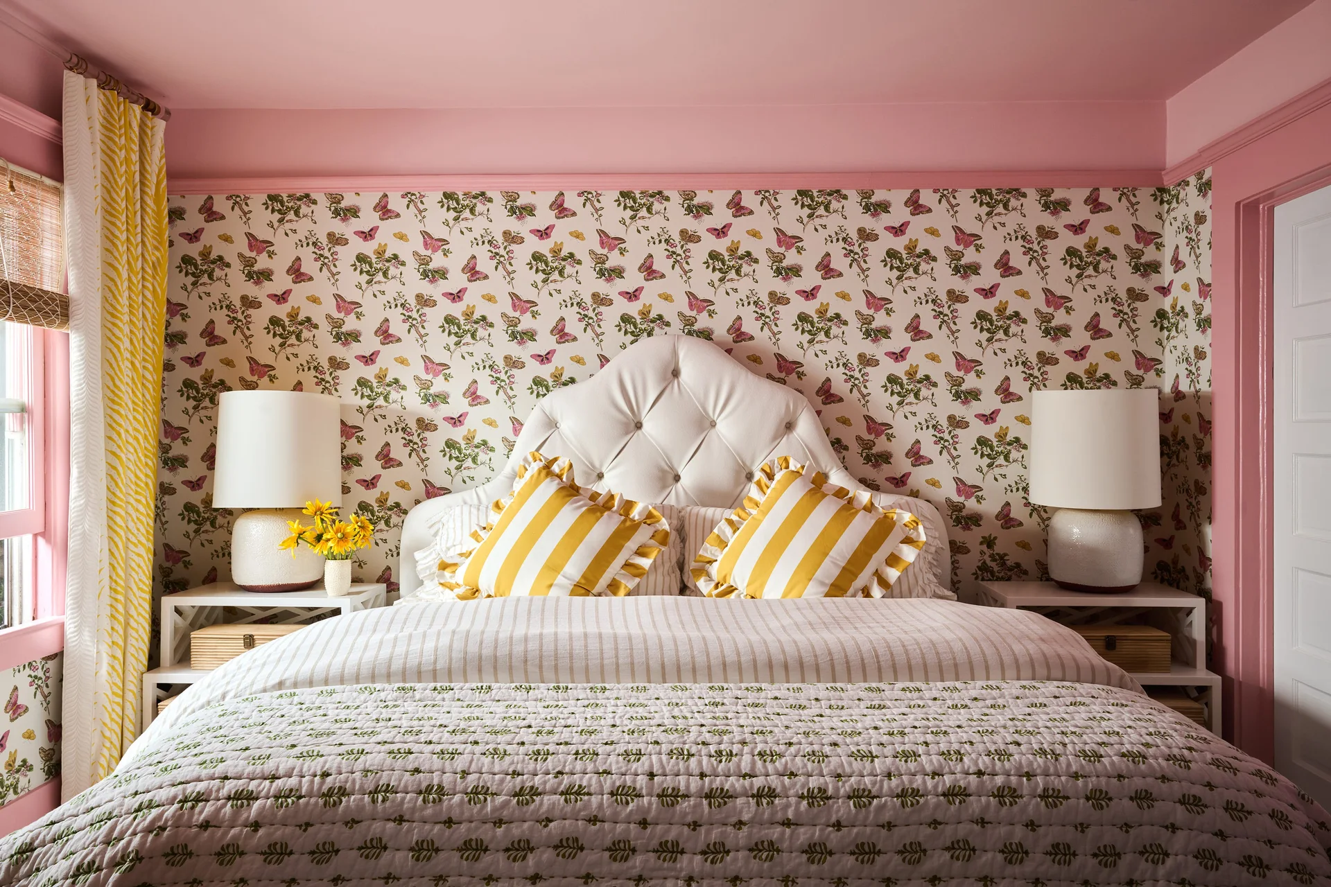A bedroom featuring a white tufted headboard, pink painted ceiling, floral butterfly wallpaper, and two yellow striped accent pillows.