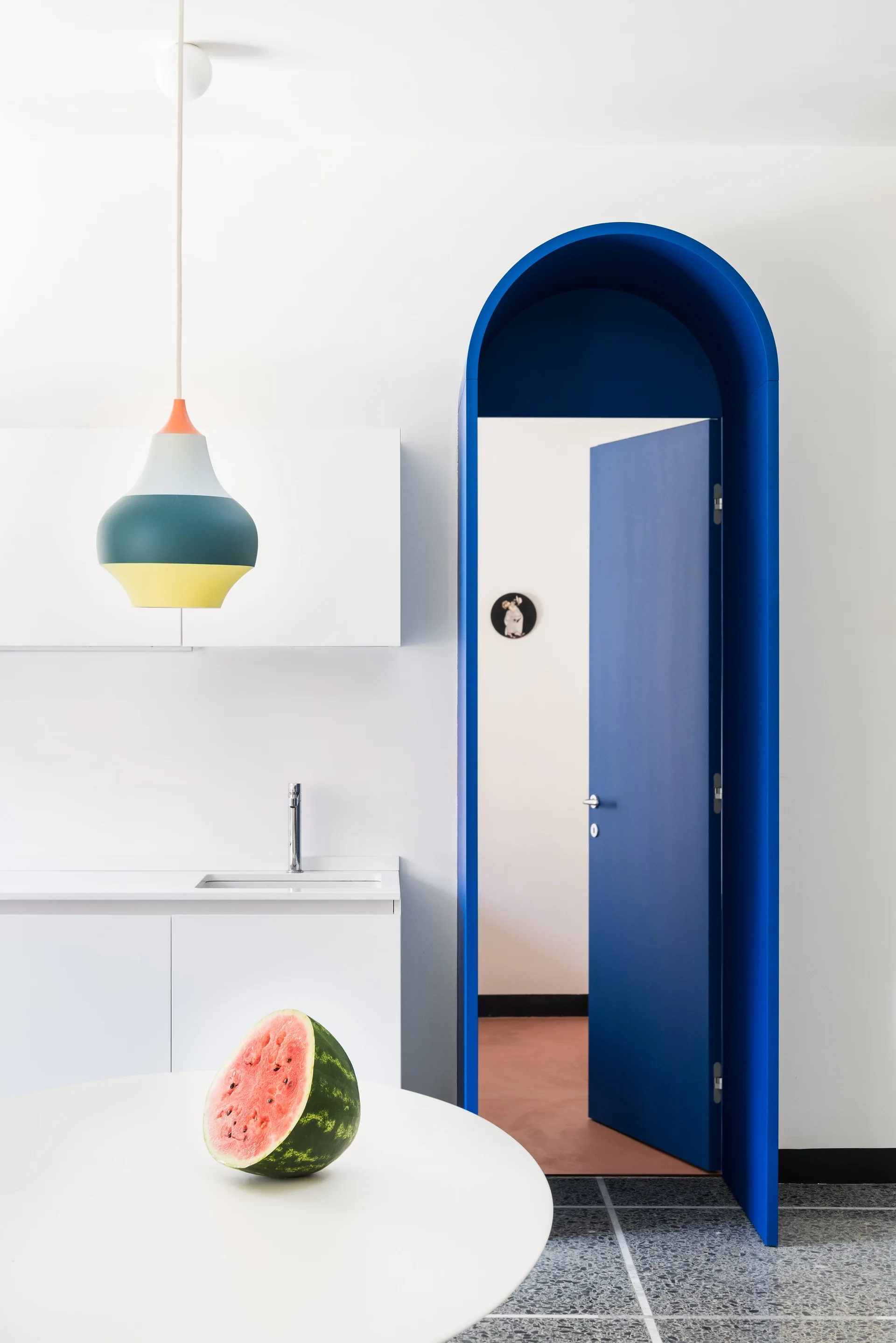 A minimalist white kitchen featuring a bold blue arched doorway, a colorful pendant light, and speckled grey terrazzo flooring.