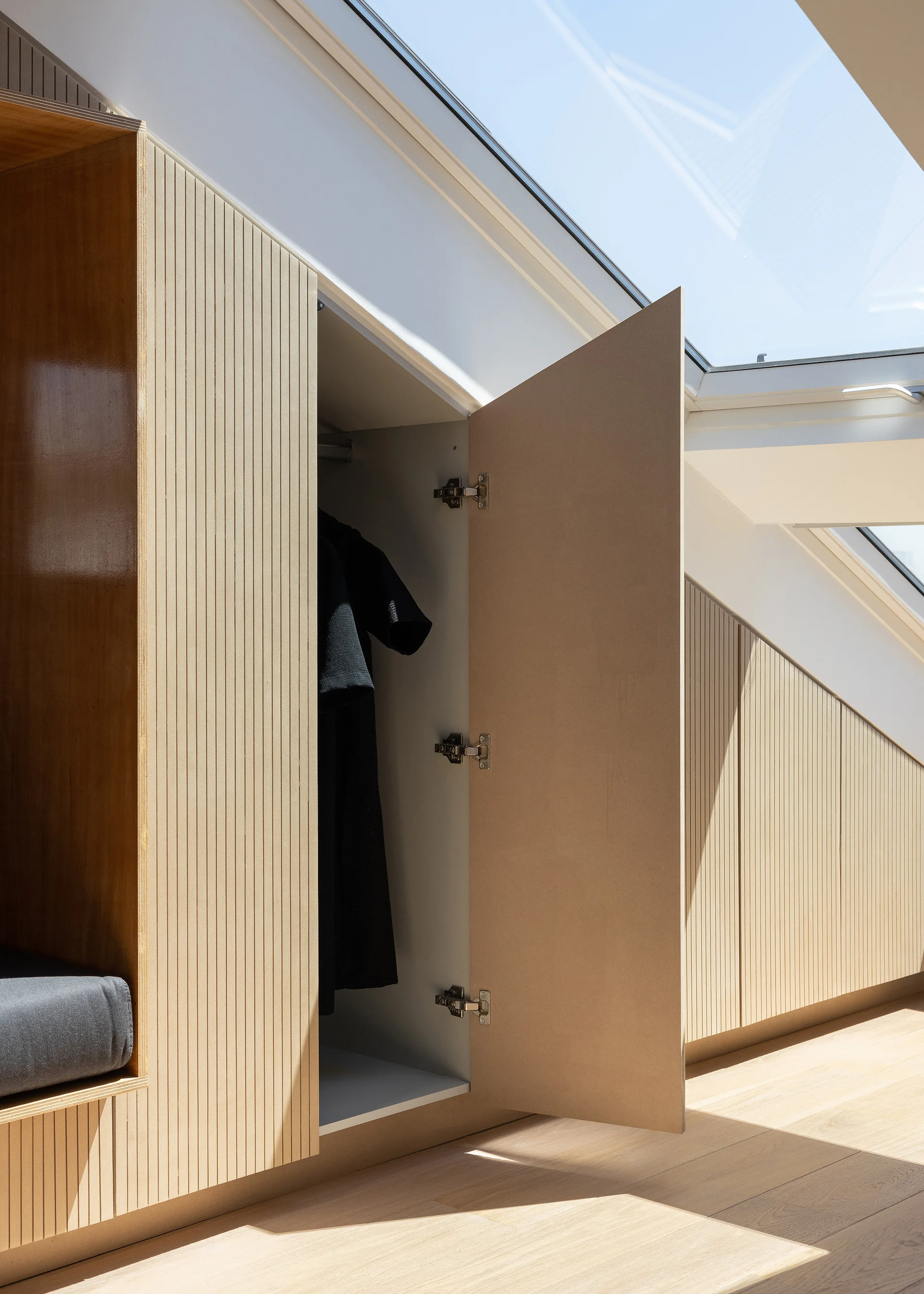 A custom light wood wardrobe with vertical slatted doors is built into a sloped attic wall under a bright skylight.