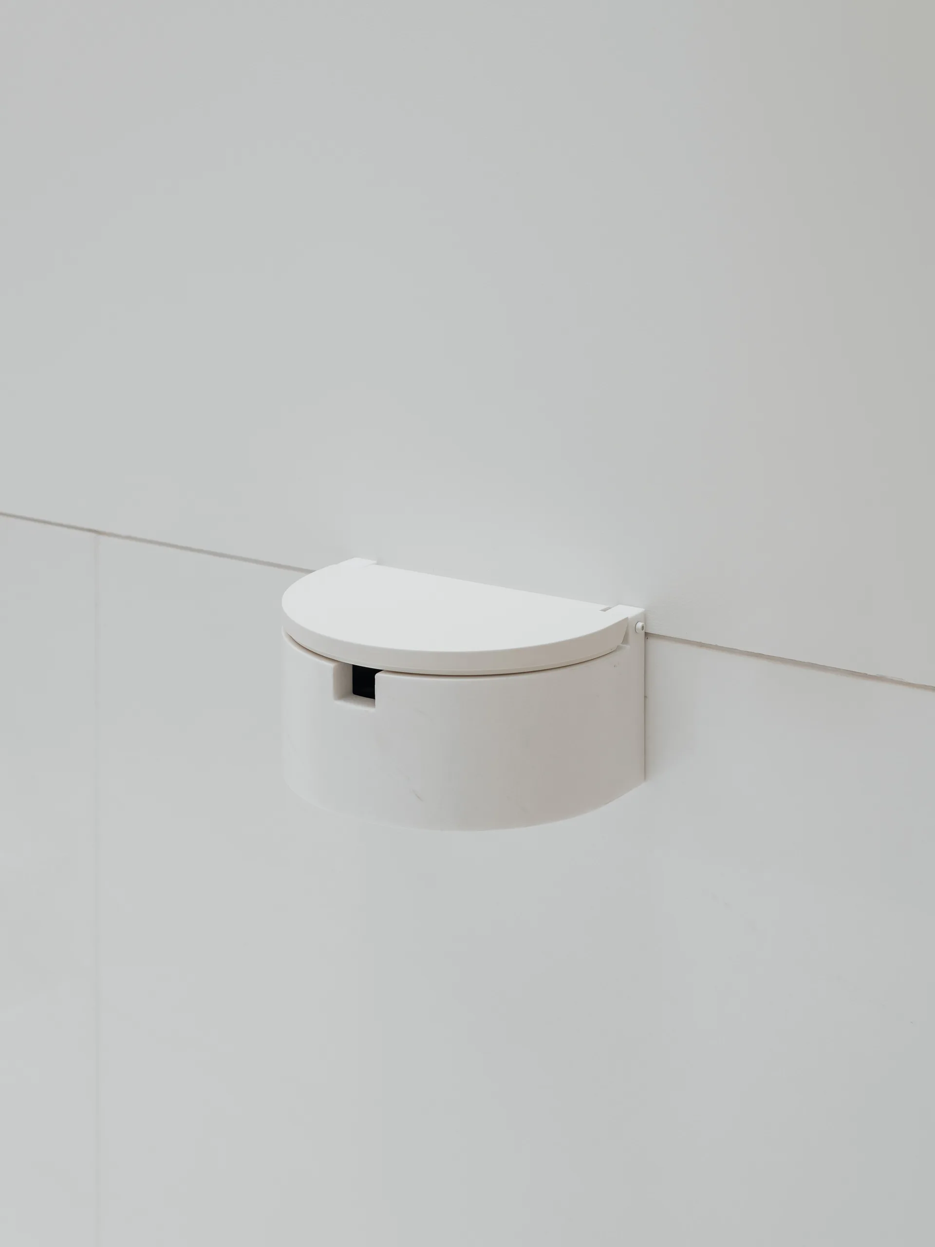 A close-up of a semicircular, matte white wall-mounted fixture with a lid, set against a smooth white tiled wall.