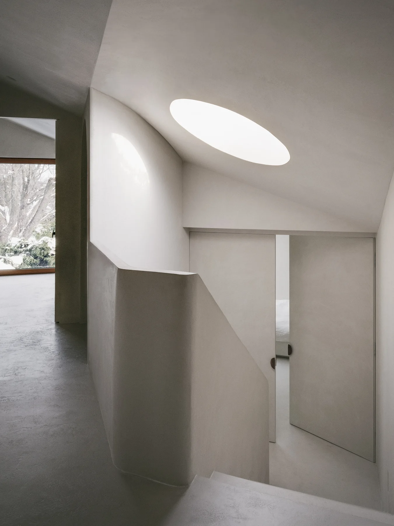 A curved plaster staircase leads down to a doorway beneath a large elliptical skylight set into a sloped ceiling.