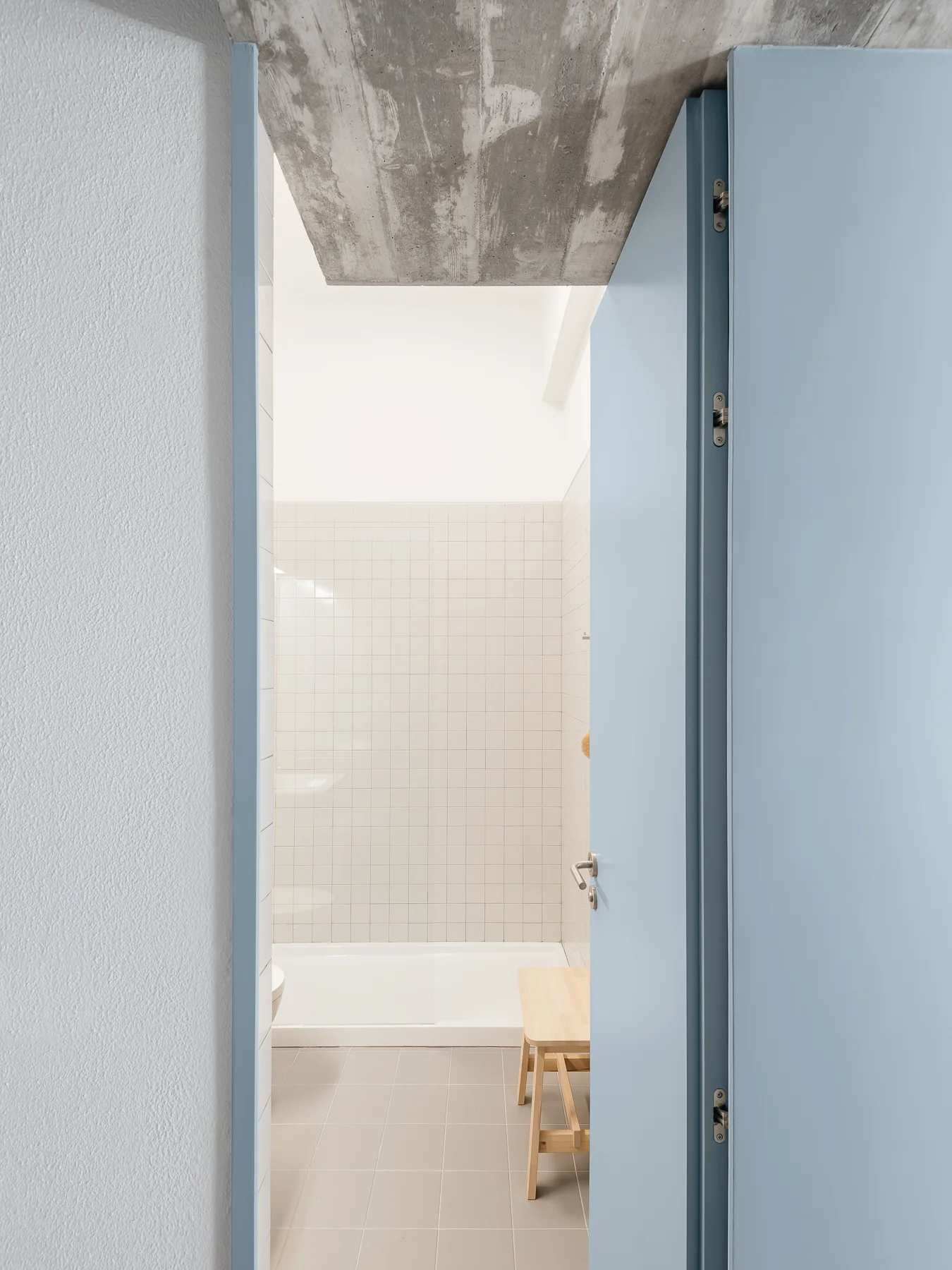 A view through a light blue doorway into a bathroom featuring white square wall tiles, a wooden stool, and an exposed concrete ceiling.