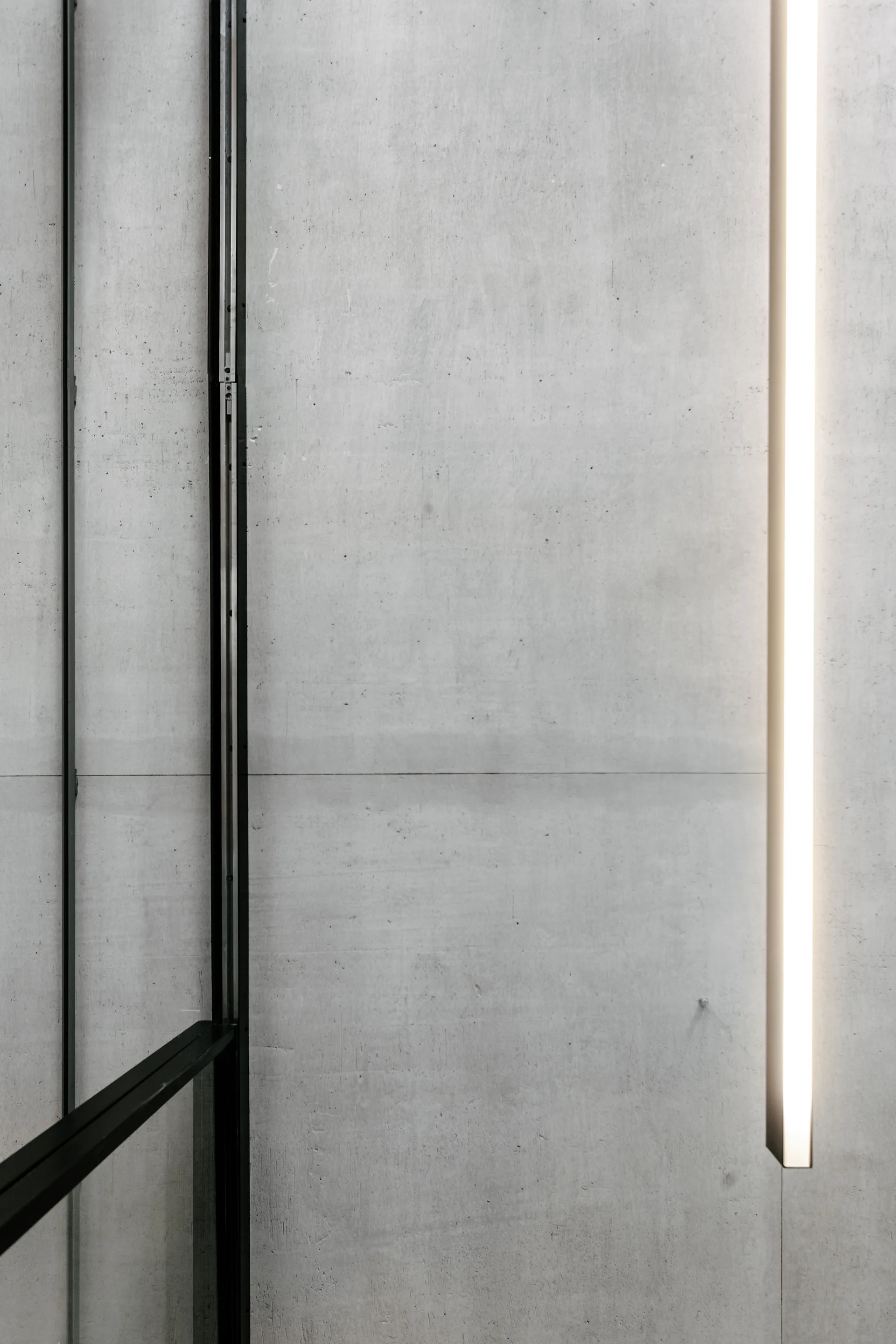 A close-up view of a grey concrete wall featuring a glowing vertical linear light fixture and a thin black metal frame.