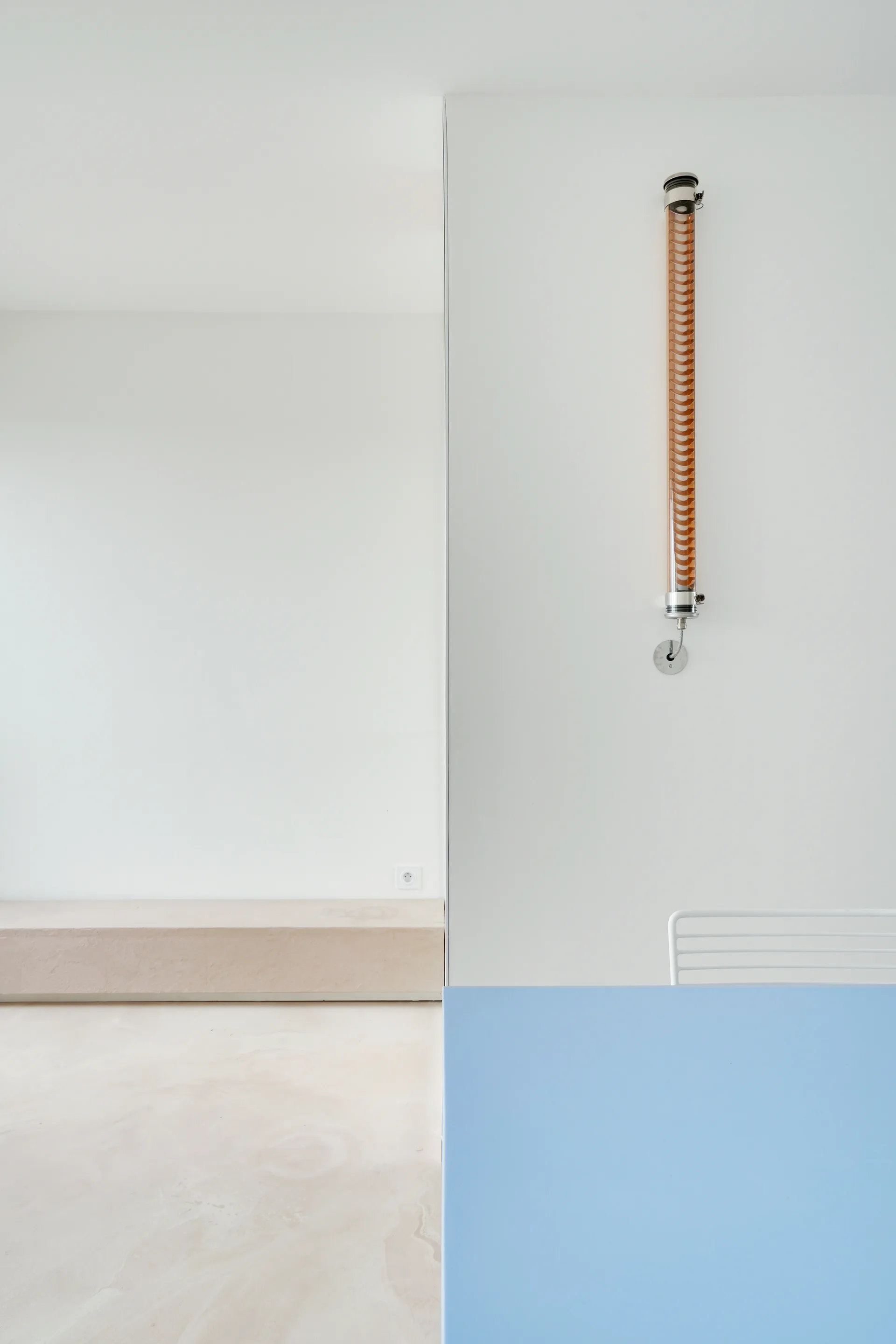 A minimalist dining area features a bright blue table in the foreground, a white metal chair, a vertical cylindrical wall sconce, and a low beige bench.