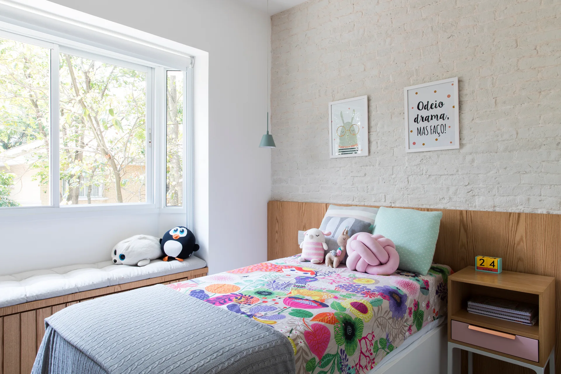 A children's bedroom featuring a white painted brick wall, a bed with colorful bedding, and a built-in wood window bench with a cushion.