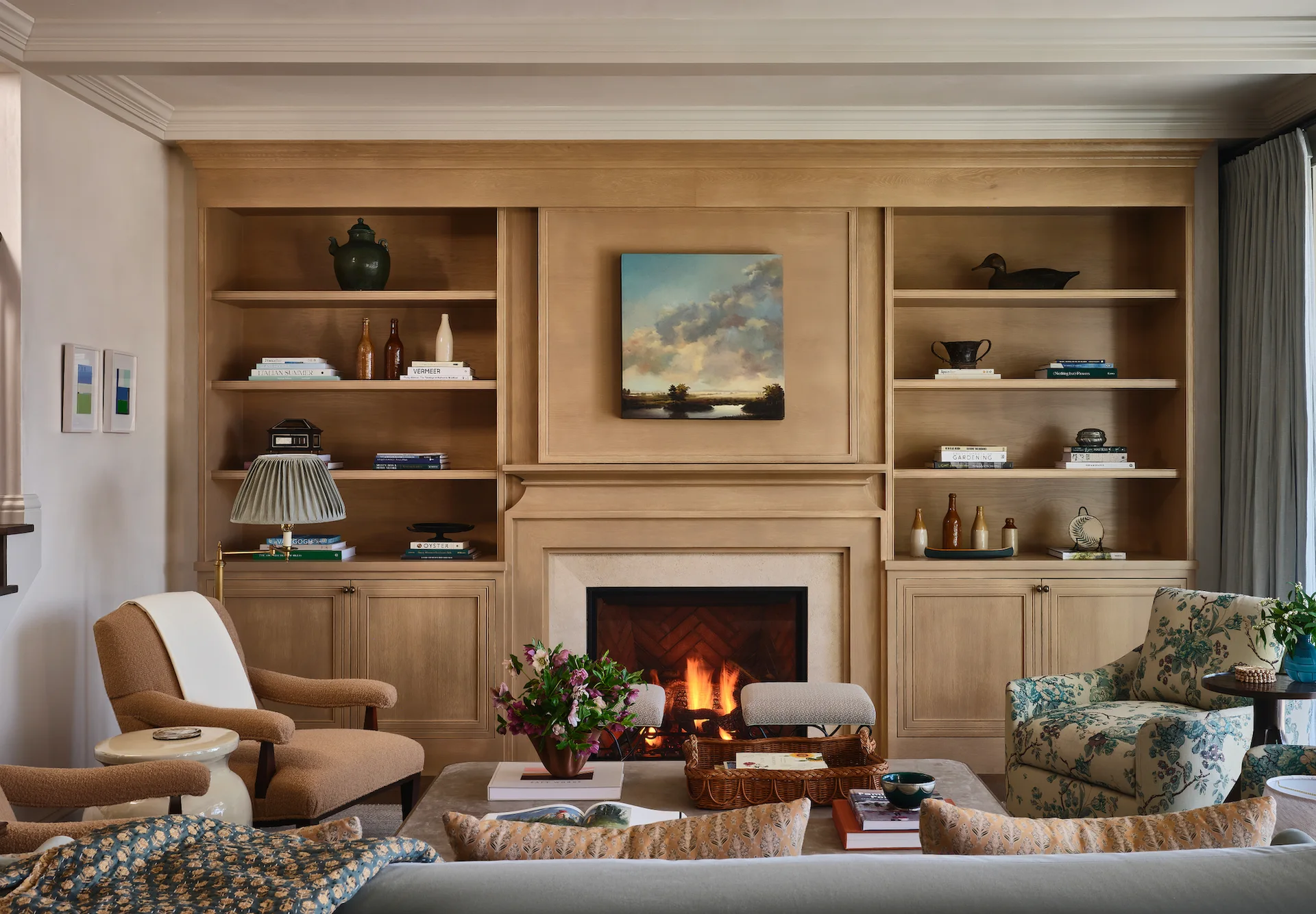 A cozy living room features a symmetrical light wood bookcase surrounding a fireplace, with two different armchairs sitting in the foreground.