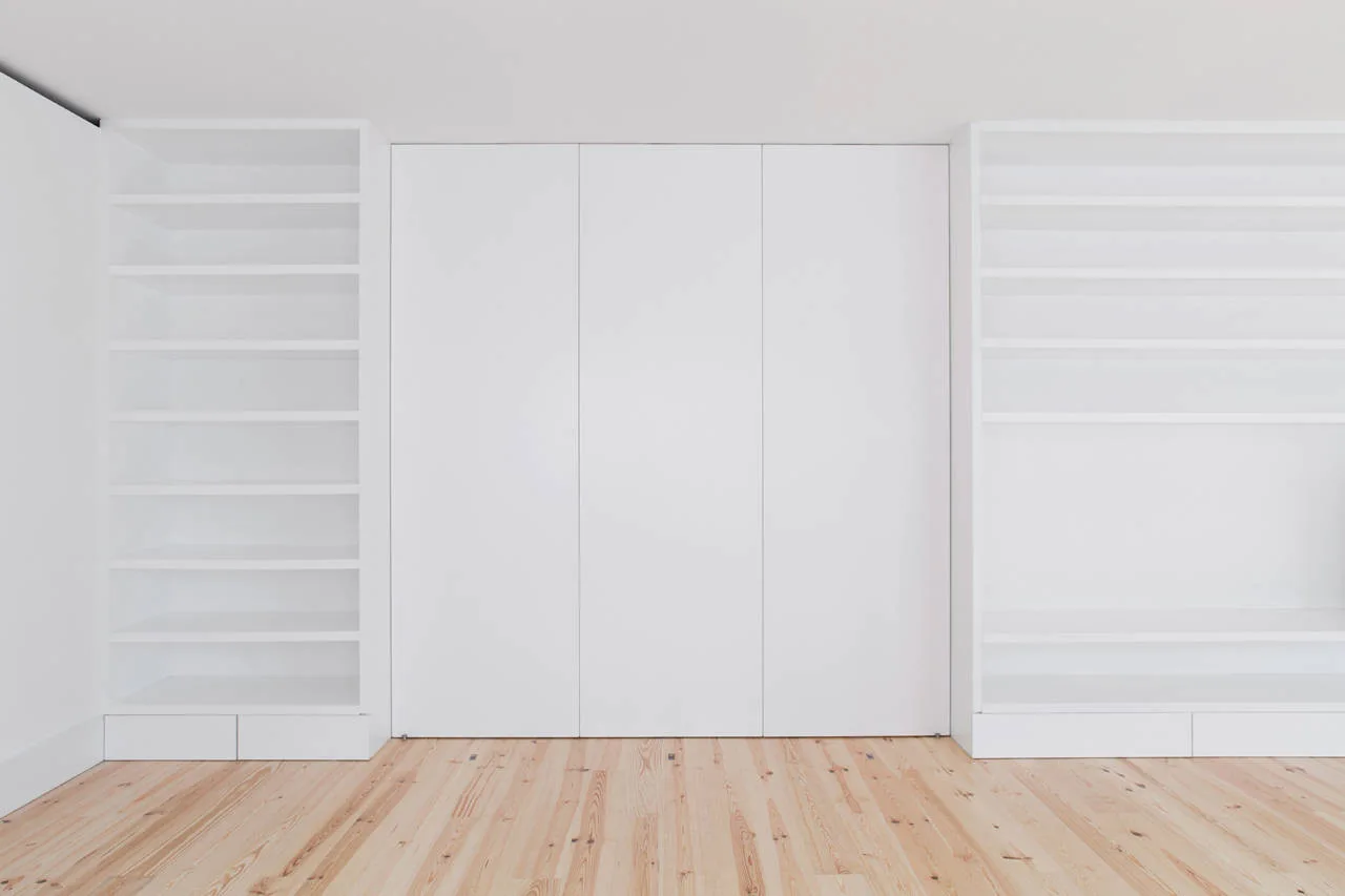 A minimalist white built-in storage unit features three flat-panel center doors flanked by empty open bookshelves over light-toned wood floorboards.