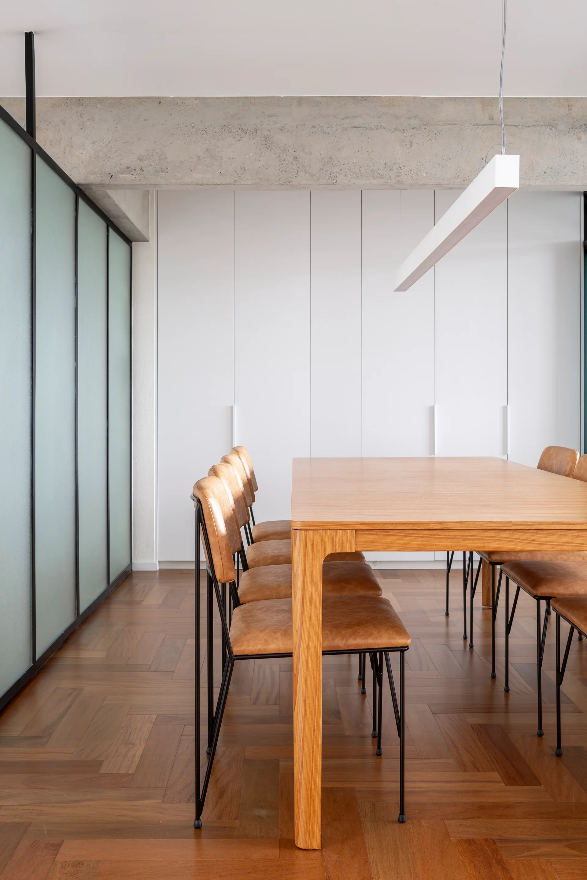 A modern dining room features a wood table with cognac chairs, set against a wall of white cabinets, an exposed concrete ceiling beam, and herringbone floors.