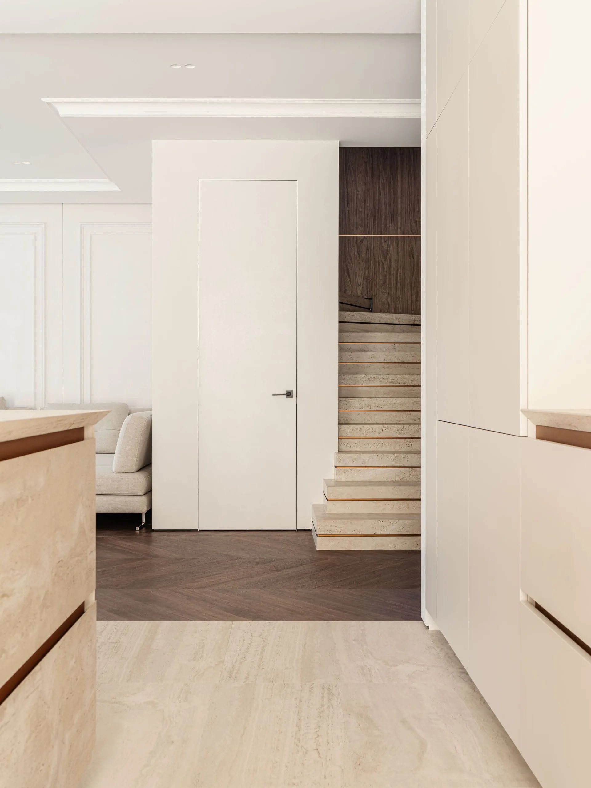 A minimalist residential interior featuring beige stone stairs next to a flush-mounted white door and dark herringbone wood flooring.