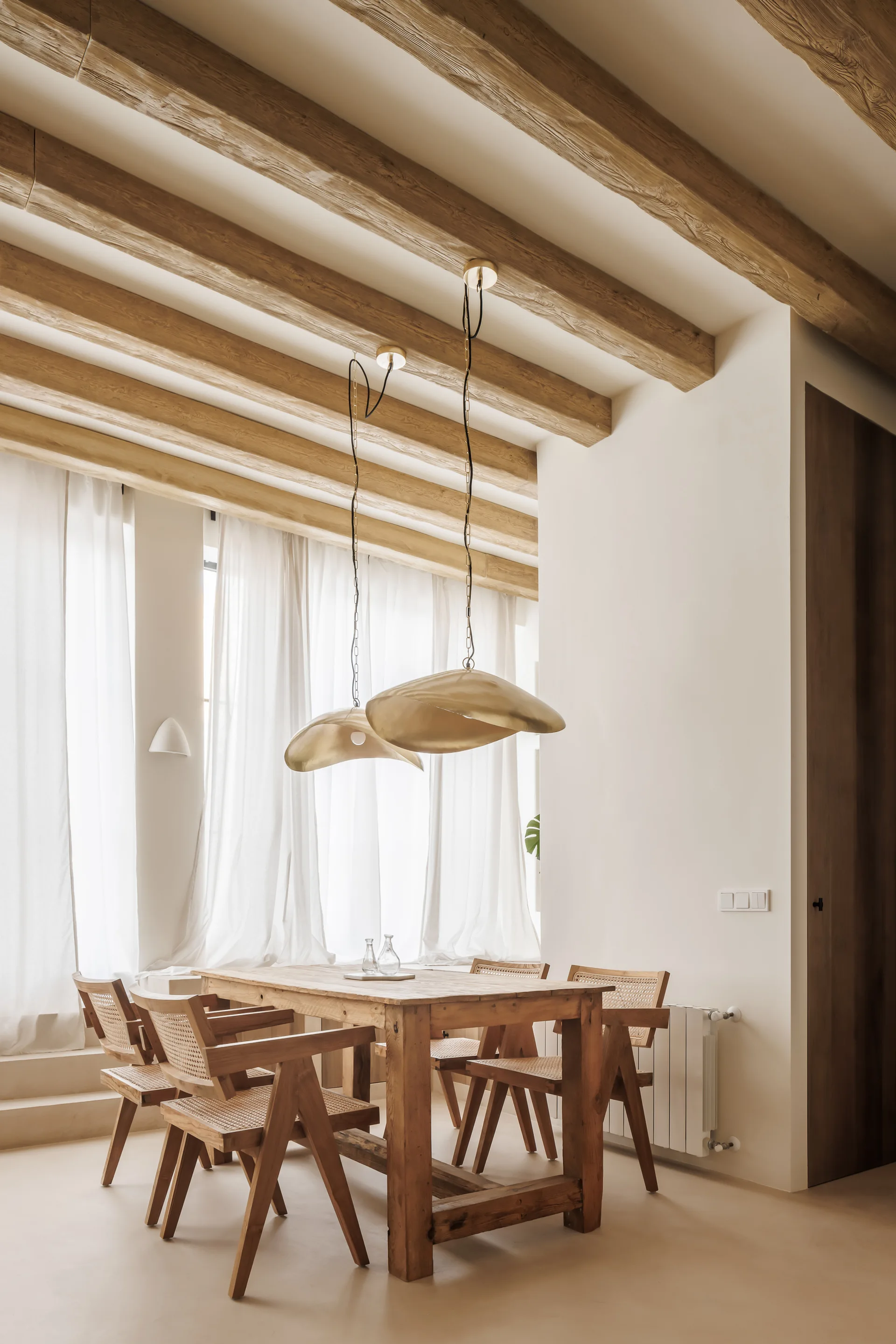 A wooden dining set sits under two brass pendant lights in a cream plaster room with exposed wood ceiling beams and sheer white curtains.