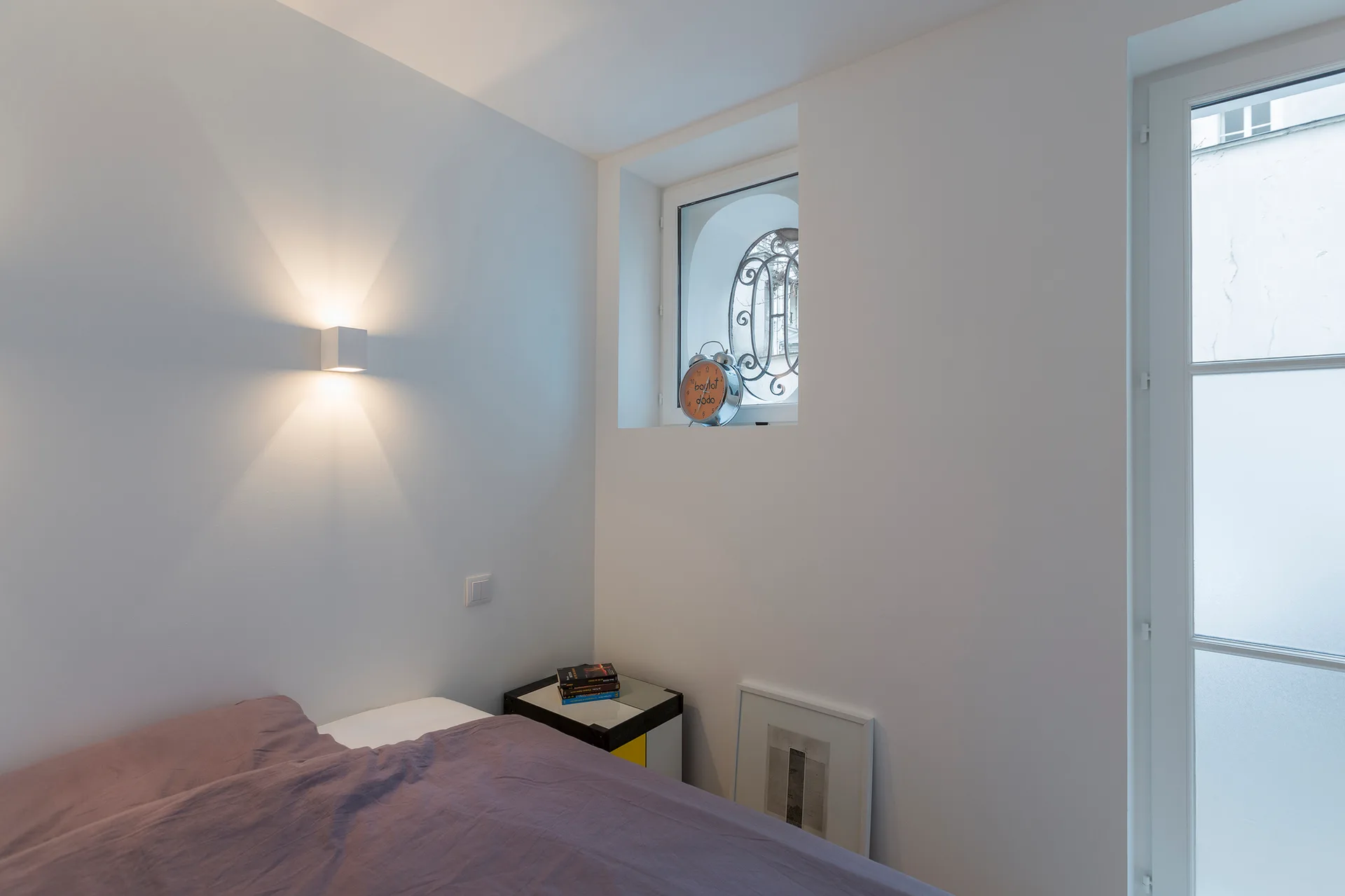 A minimalist white bedroom features a bed with purple bedding, a small square nightstand, a wall sconce, and a recessed window.