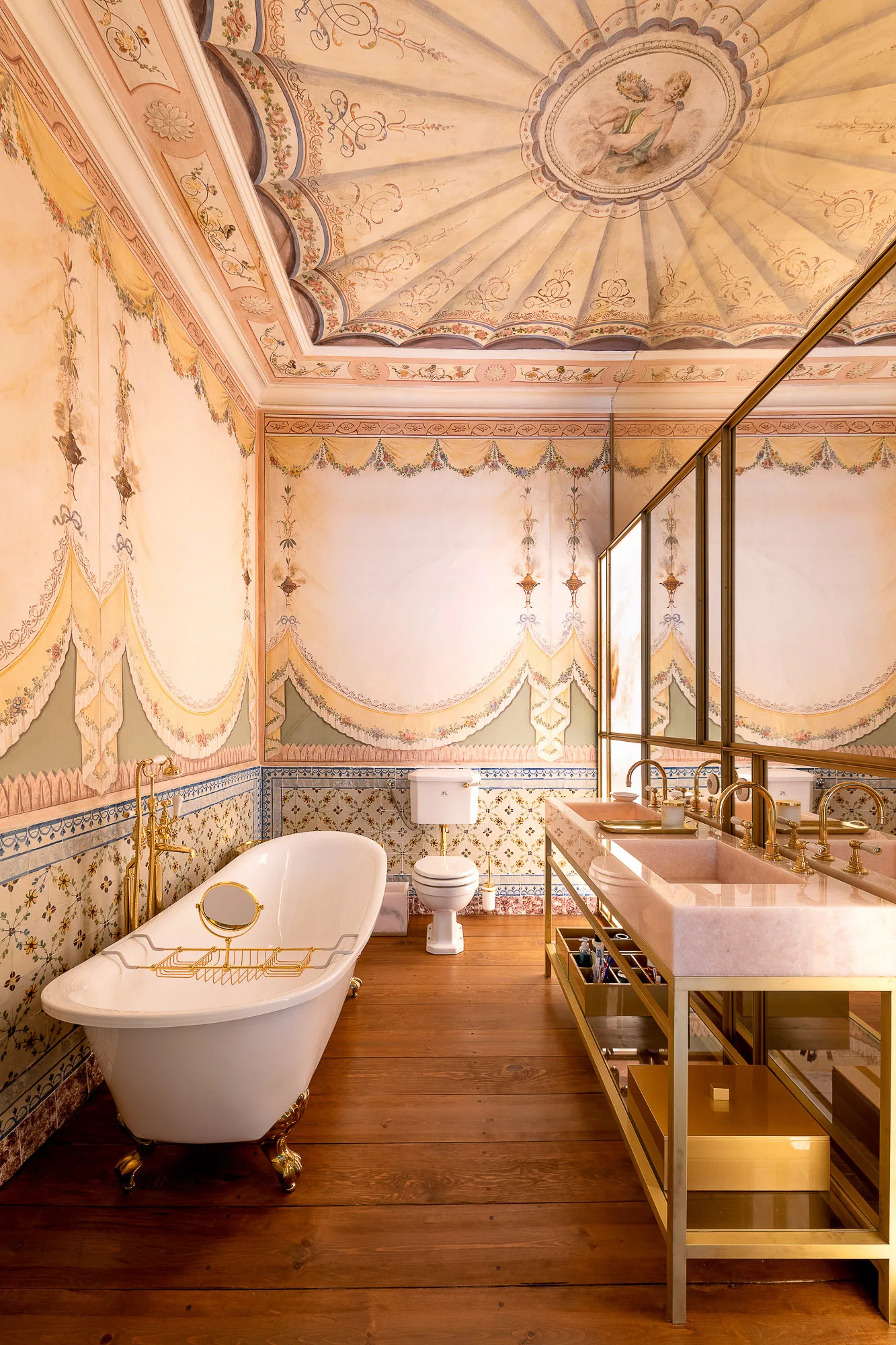 A luxury bathroom features a hand-painted mural ceiling, a pink stone double vanity with brass fixtures, and a freestanding white clawfoot tub.
